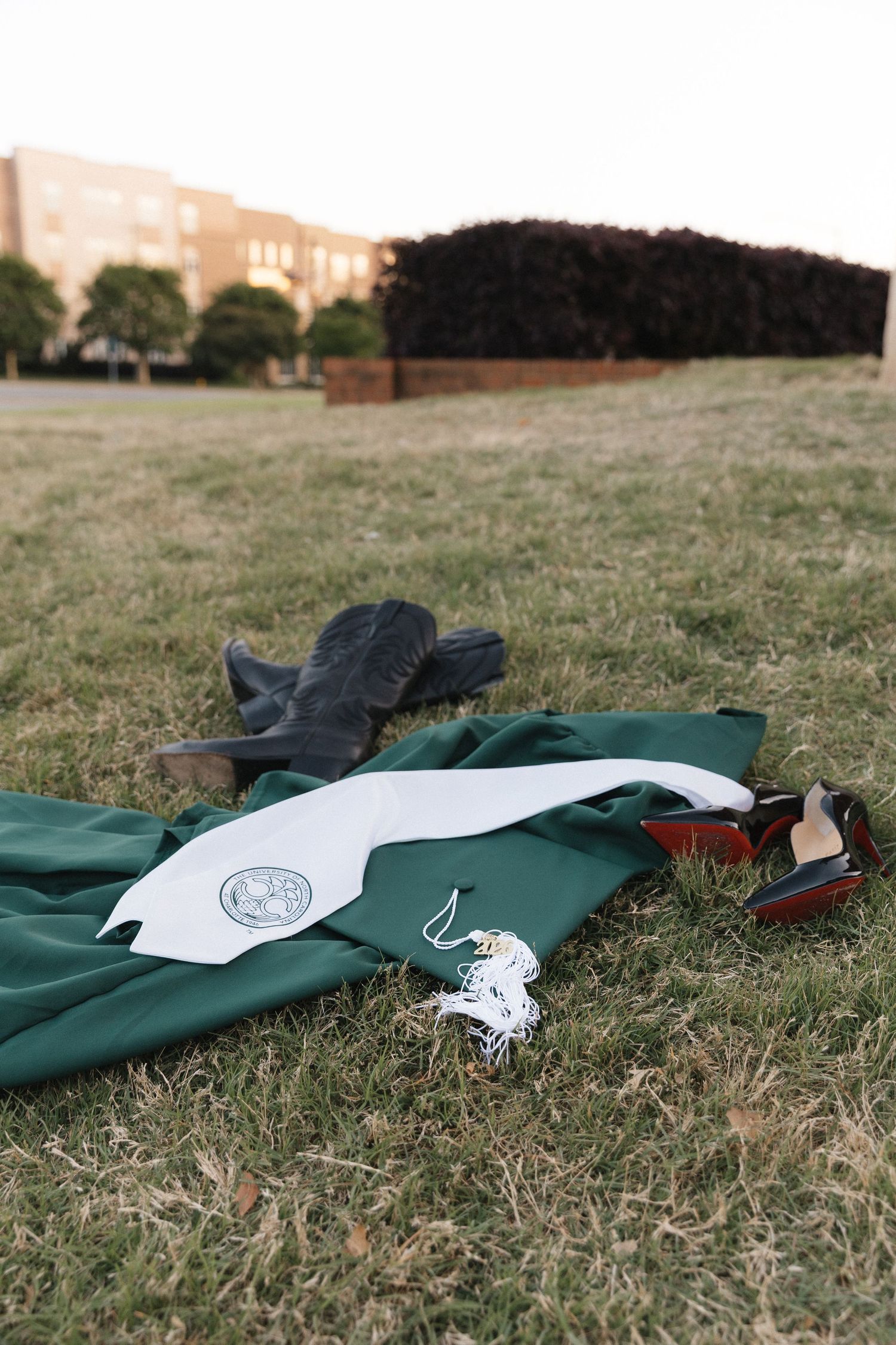 Green graduation gown, white stole with seal, cap, tassel, and sunglasses laid on grass near university building.