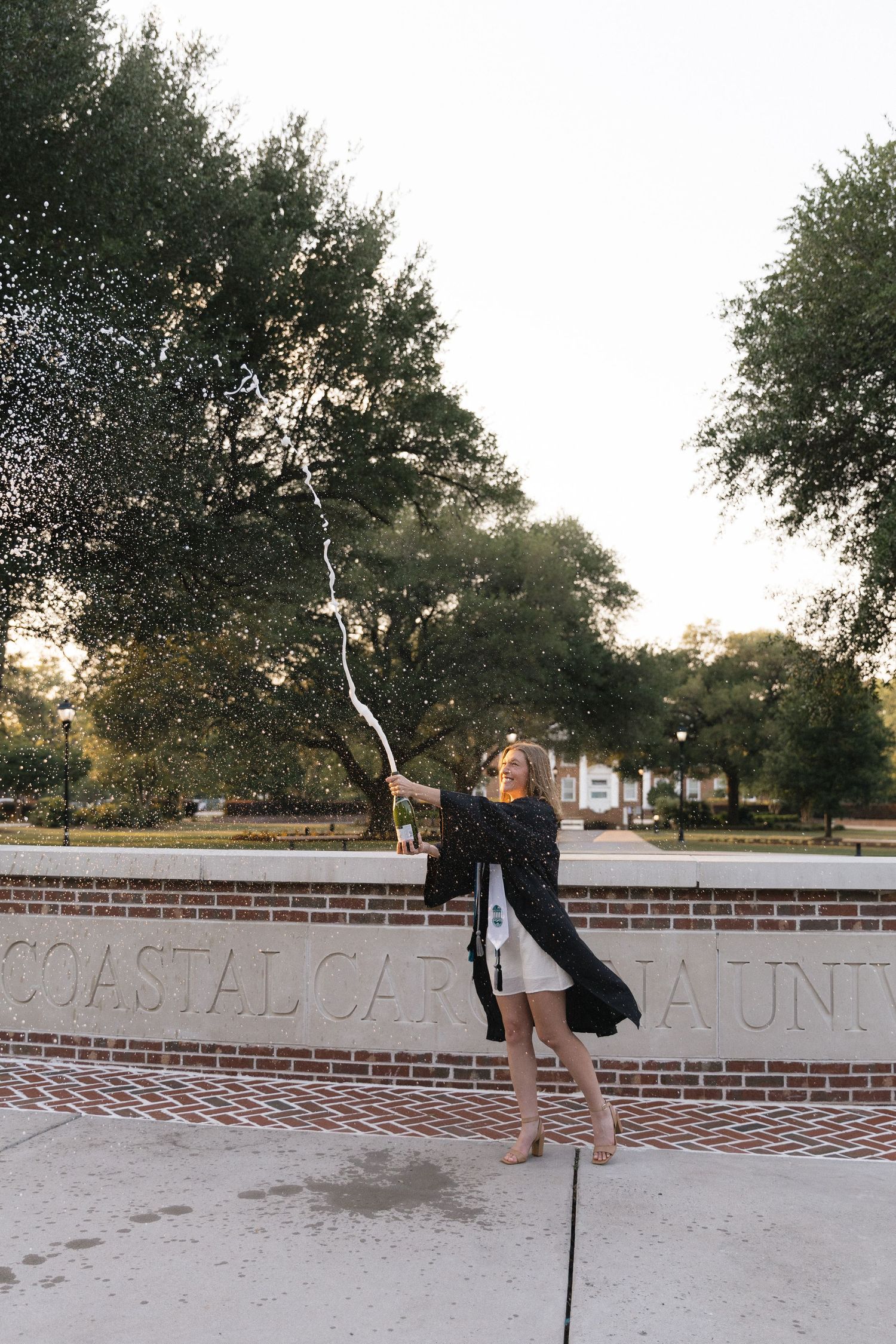 Graduate in cap and gown celebrates with sparkler at Coastal Carolina University brick entrance sign.