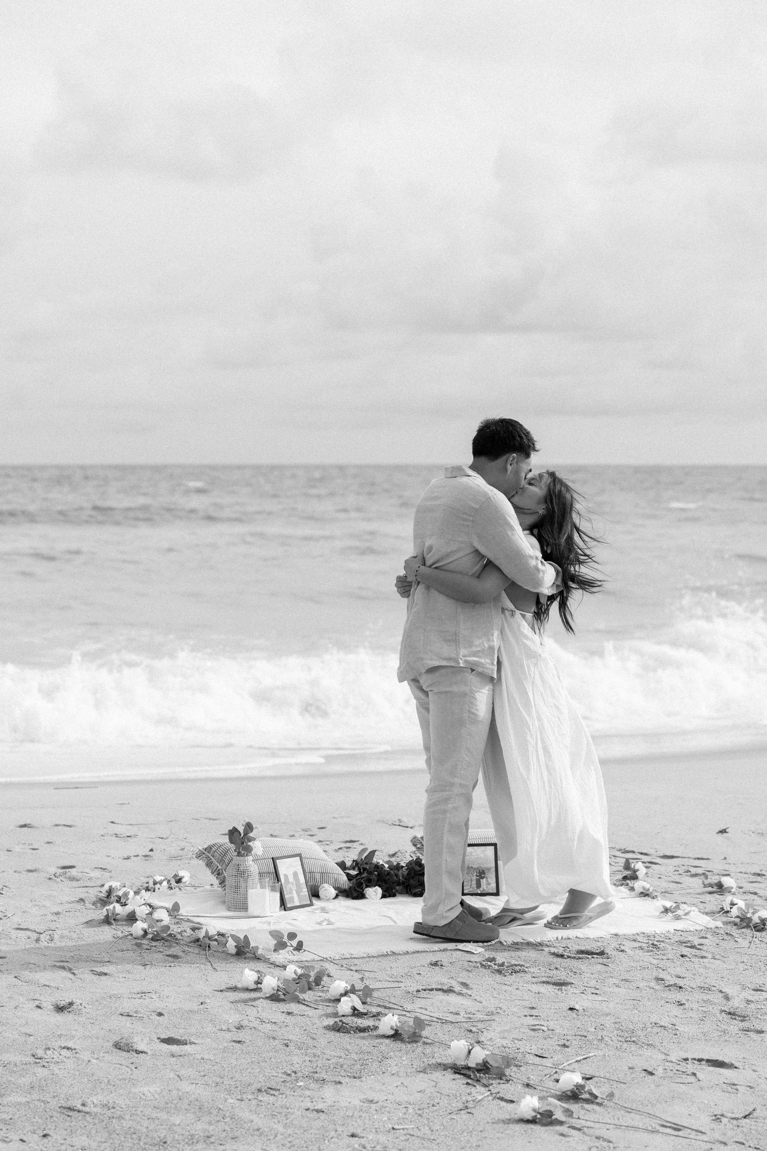 Black and white photo of a couple embracing on a beach near the ocean, surrounded by flowers and framed photos on sand.
