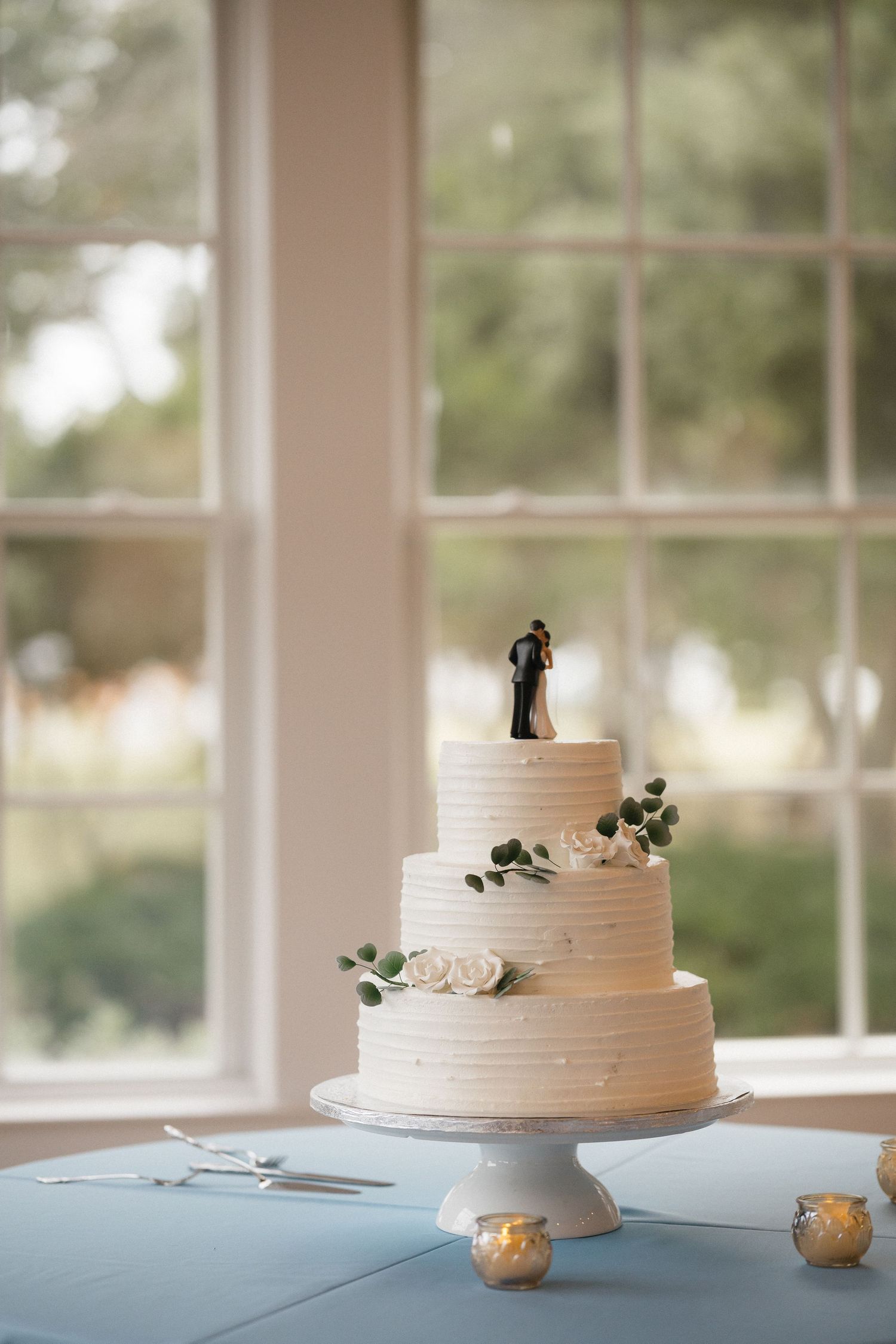 Three-tier white wedding cake with bride and groom topper, white roses, and greenery on a blue tablecloth near a window.