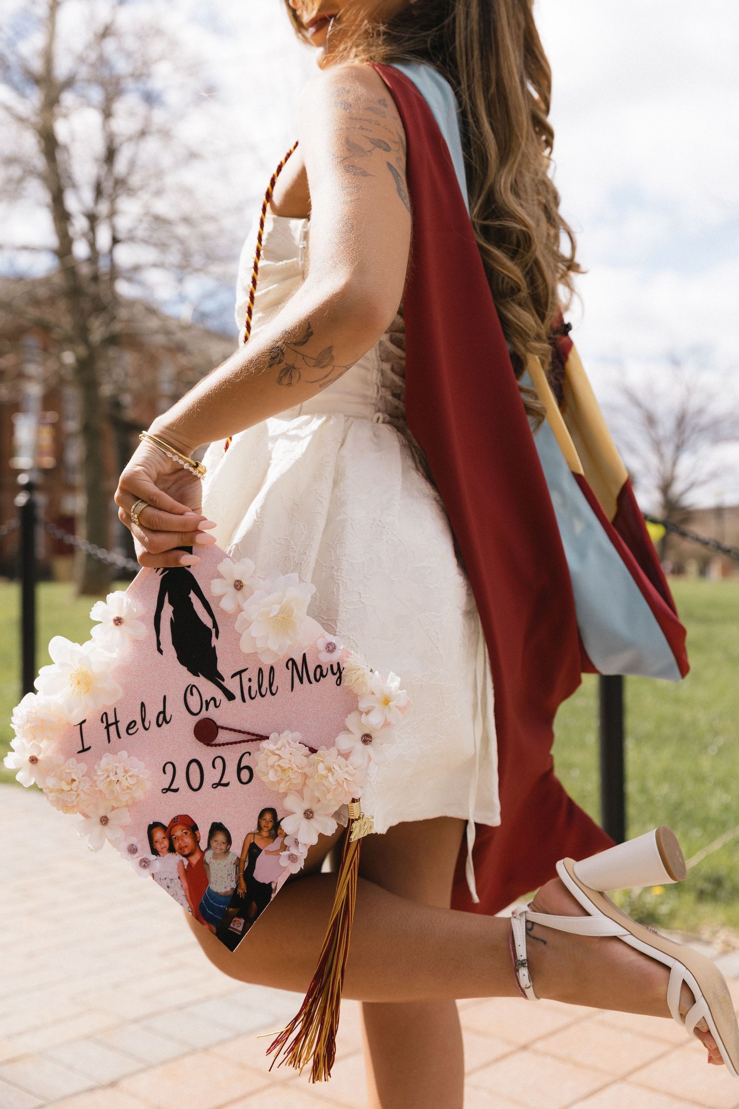 Graduate holds a decorated cap reading I Held On Till May 2026 while wearing a white dress and maroon regalia.