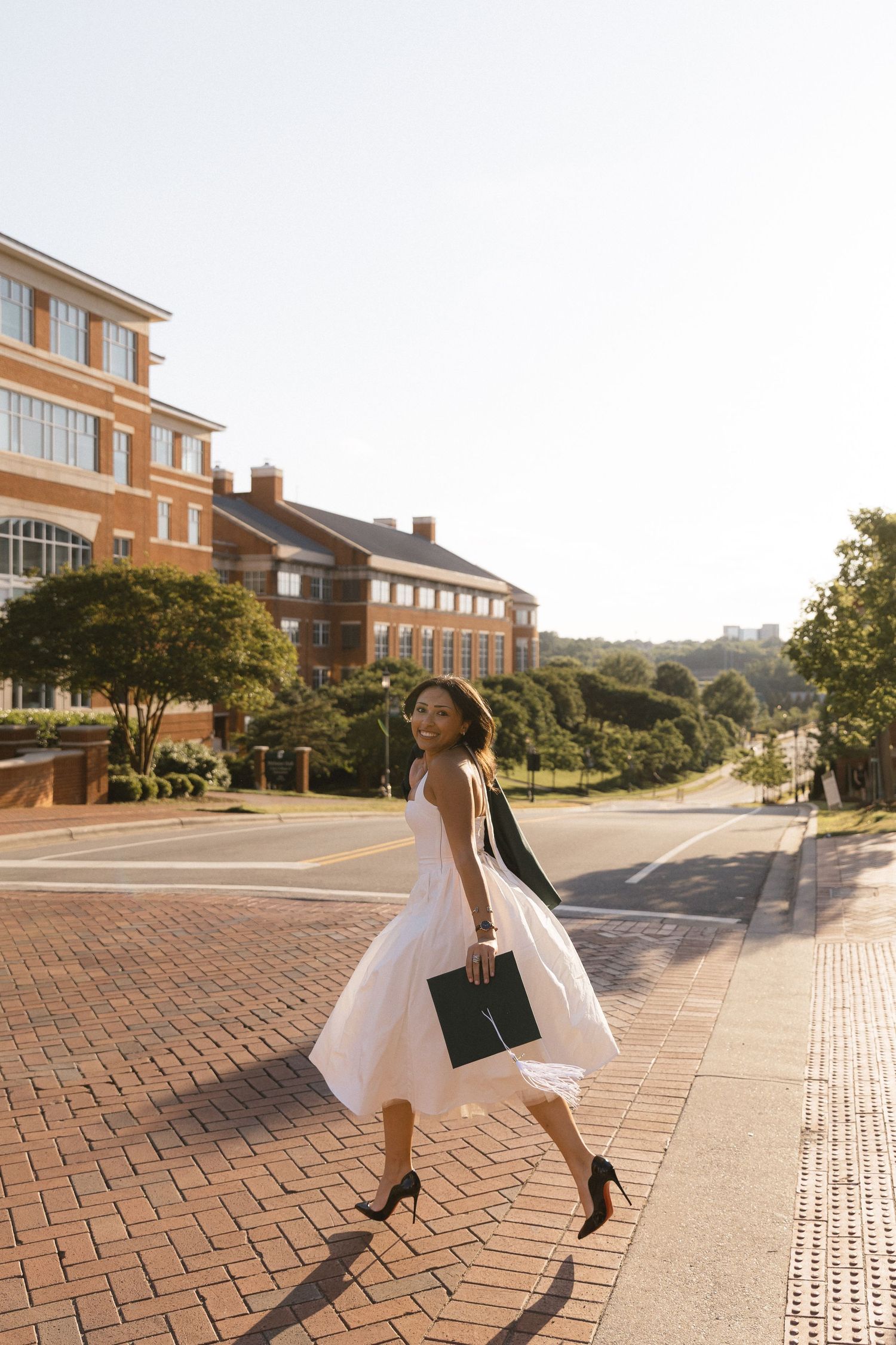 Young woman in white midi dress holding graduation cap walks across brick plaza near red brick university buildings.
