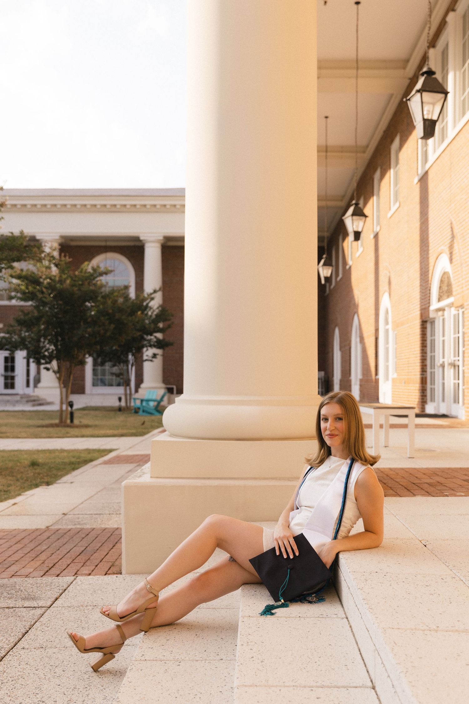 Young woman in white top and black skirt sitting against a column on a university campus building exterior.