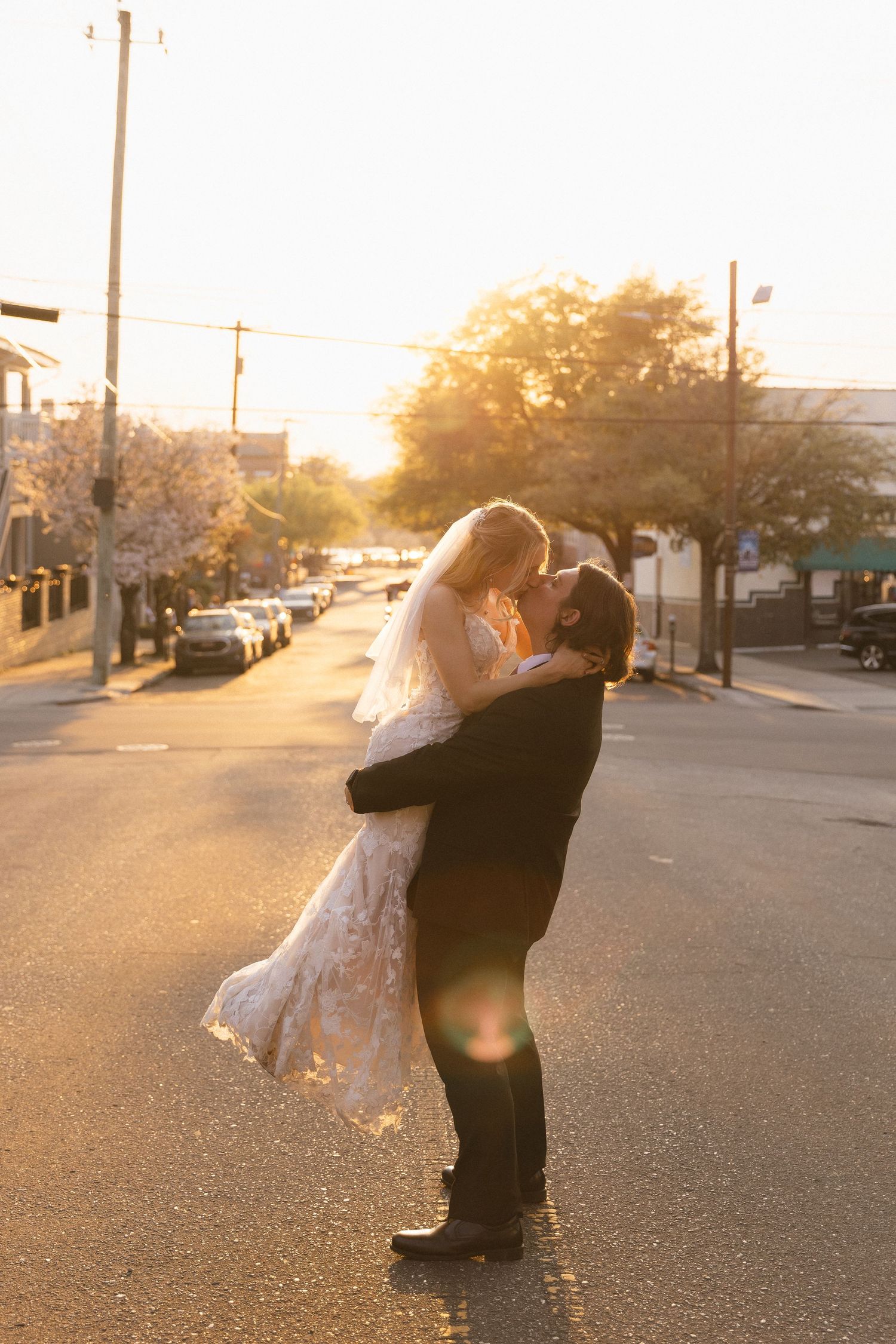 Groom lifts bride in wedding attire on sunlit street, sharing a romantic kiss with golden hour light and cherry blossoms.