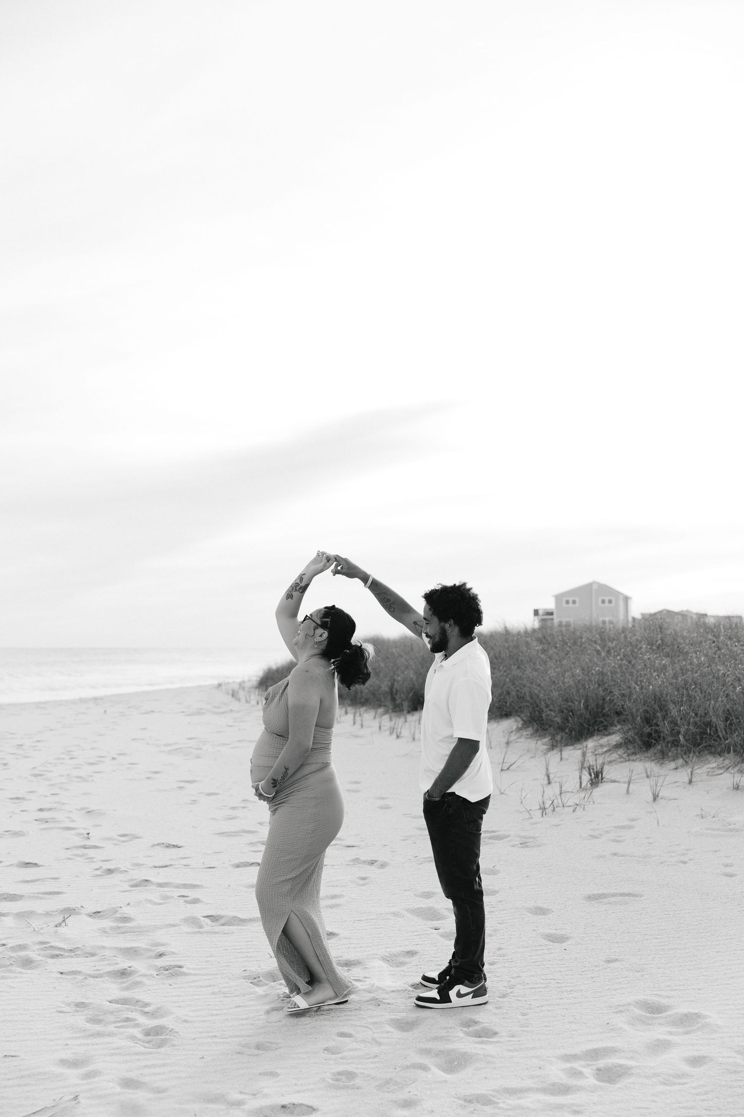 Black and white photo of a couple dancing on a sandy beach, woman in flowing dress being twirled by man near dunes.