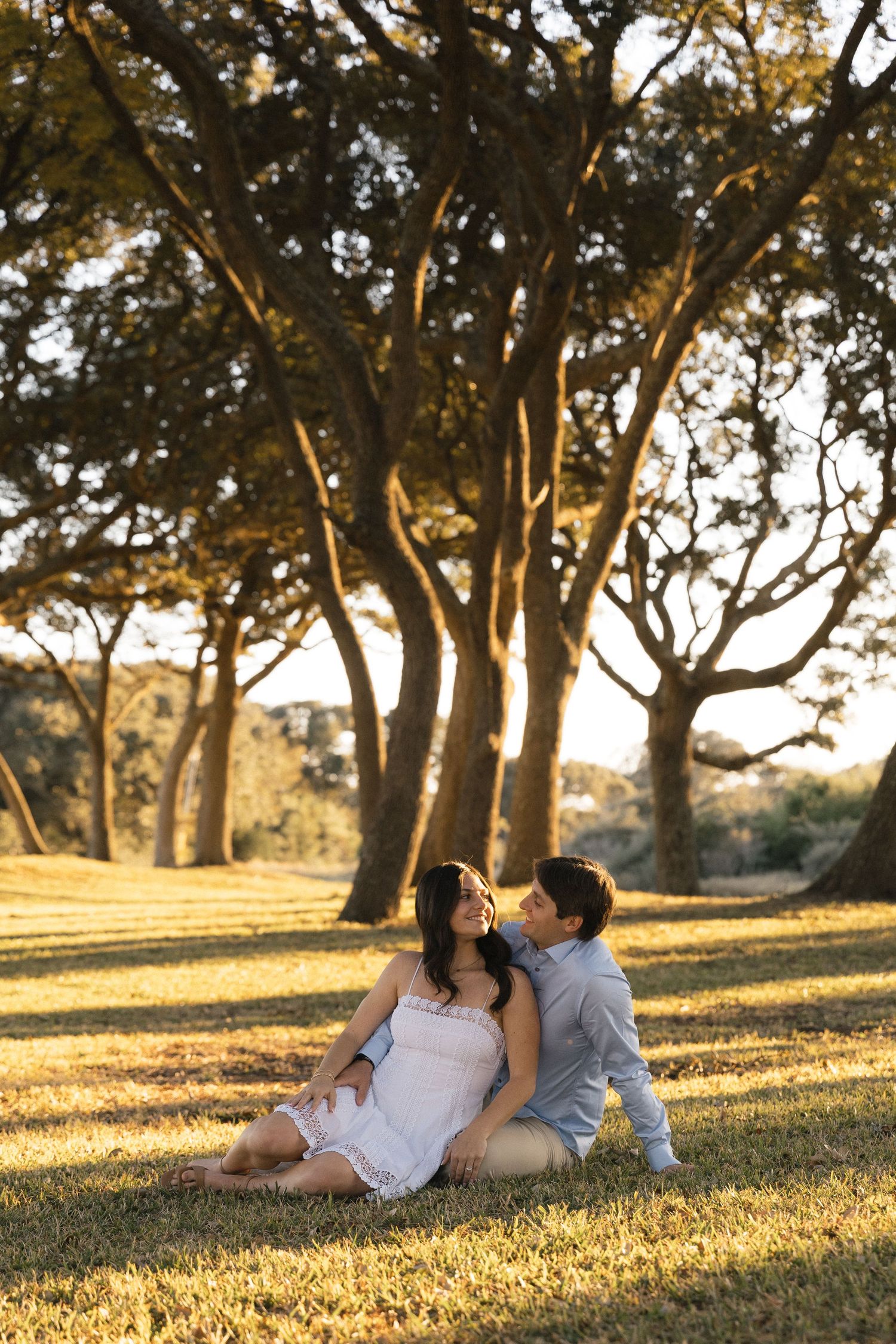 Couple sitting on grass in golden hour sunlight, leaning together romantically beneath large oak trees in a park.