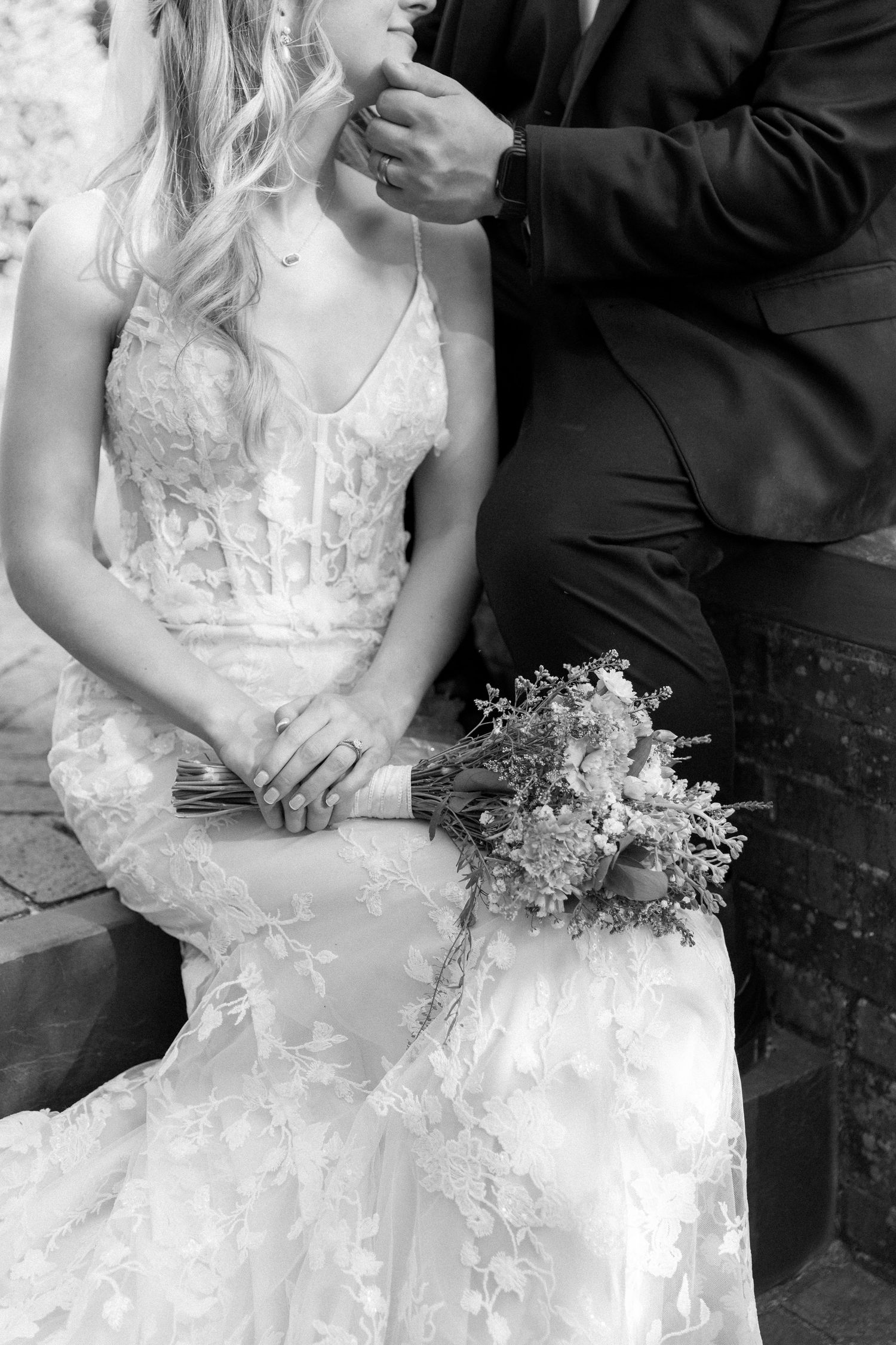 Black and white wedding photo of bride in lace gown holding bouquet while groom adjusts her hair.