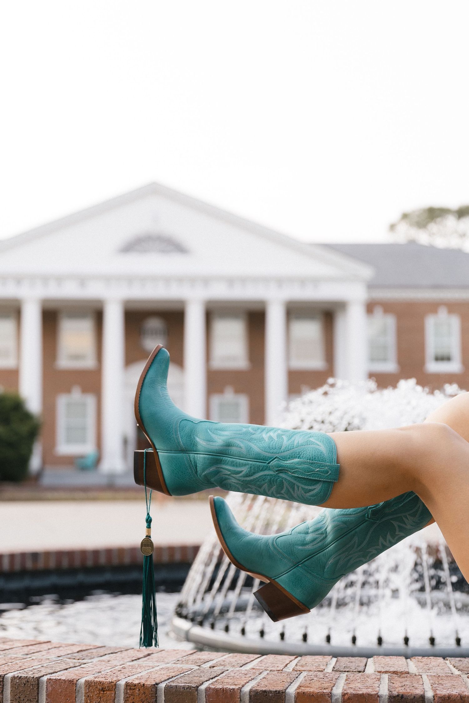 Teal cowboy boots with tassel shown near a brick fountain outside a classic brick building.