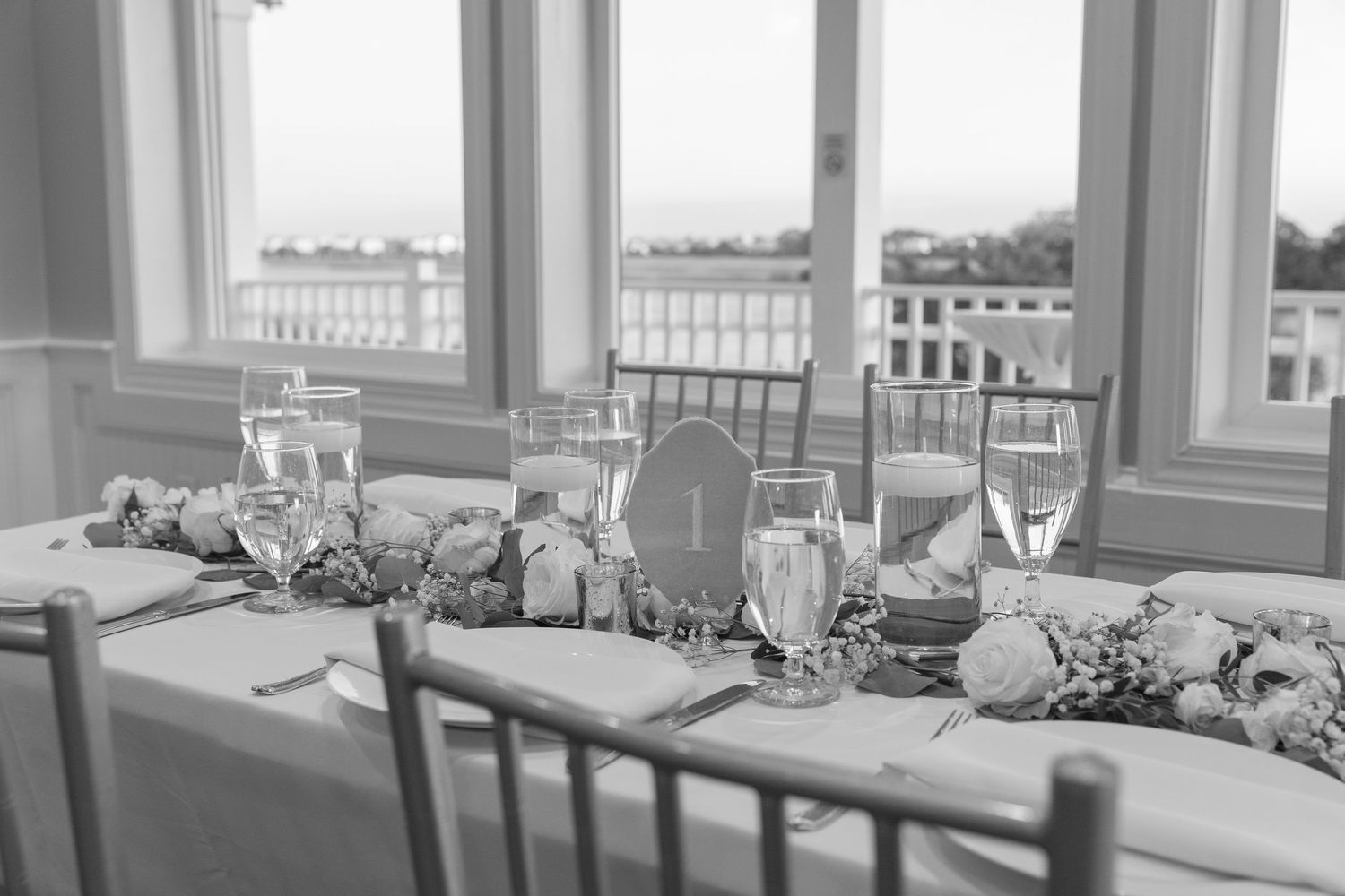 Elegant black and white wedding reception table with floral centerpieces, candles, and chiavari chairs near large windows.