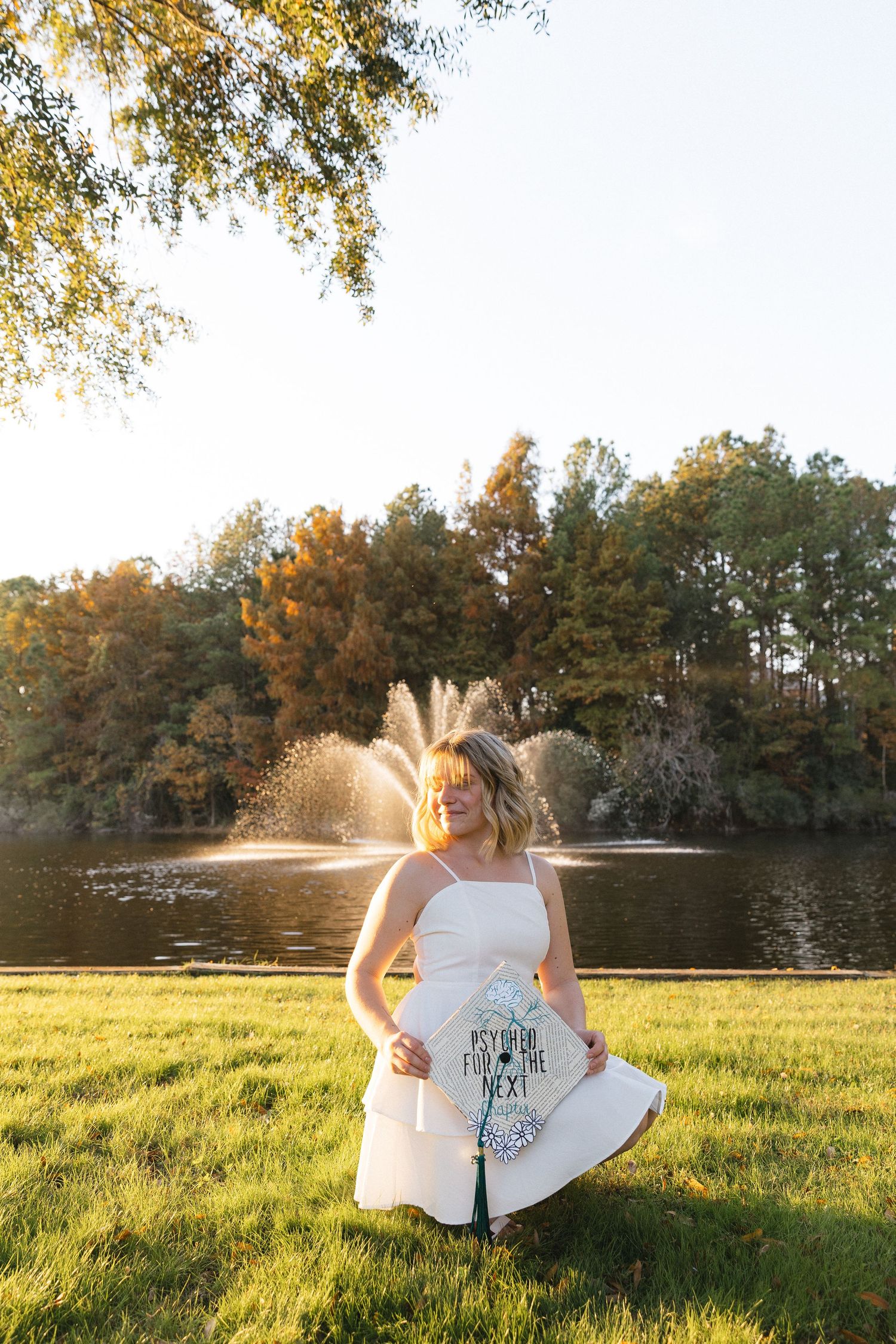 Woman in white dress holding graduation cap stands by a sunlit pond with fountain and autumn trees.