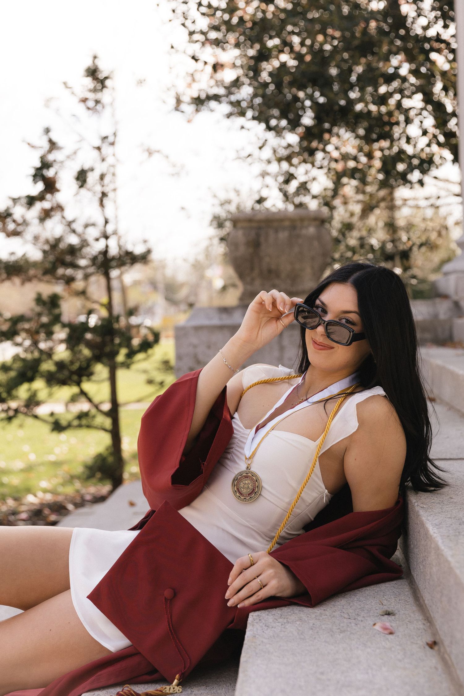 A young woman in a white dress and maroon graduation gown poses on steps, wearing sunglasses and a gold medal.