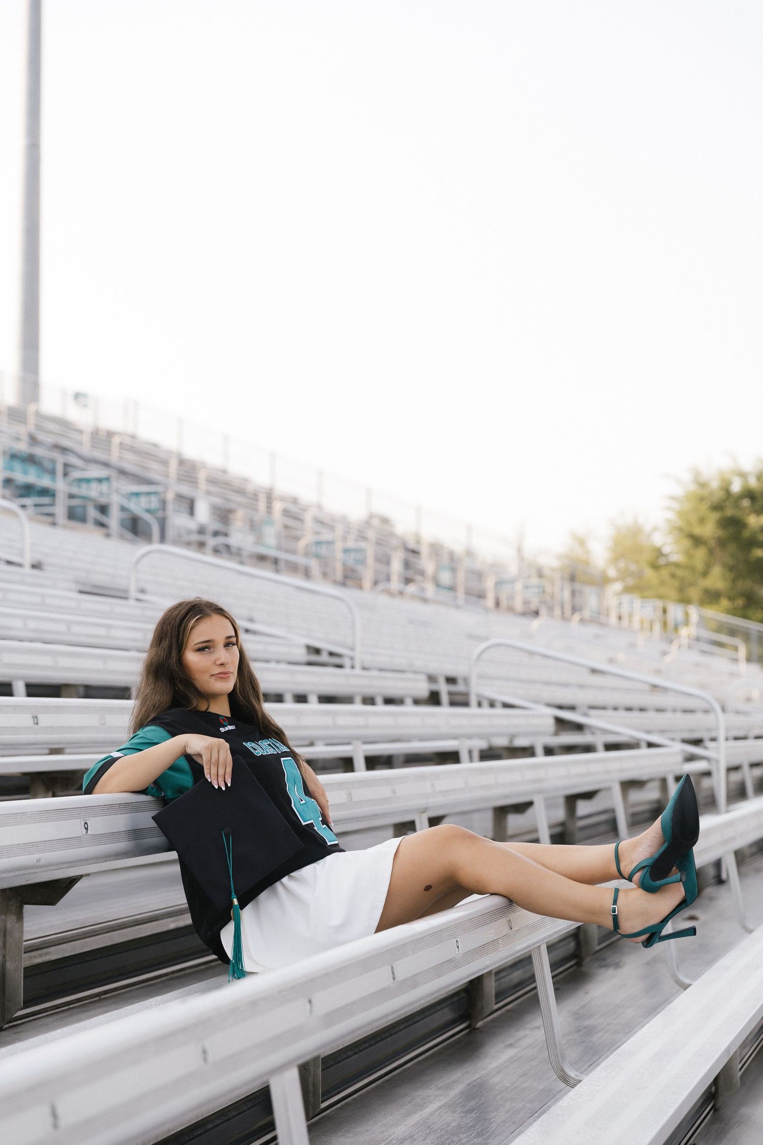Young woman in black jersey and white skirt poses on bleachers wearing teal heels.