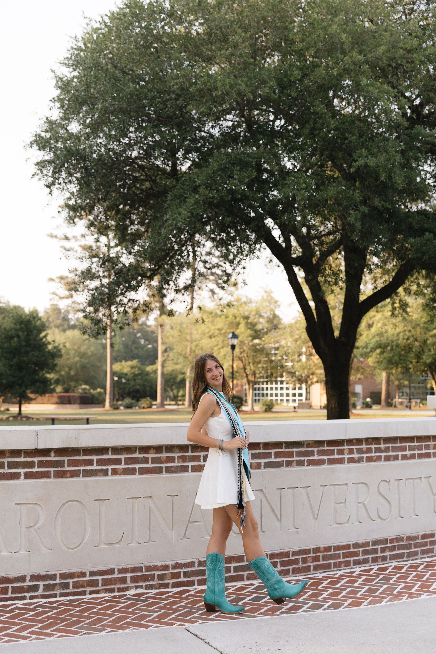 Young woman in white dress and teal cowboy boots poses by Carolina University brick sign outdoors.