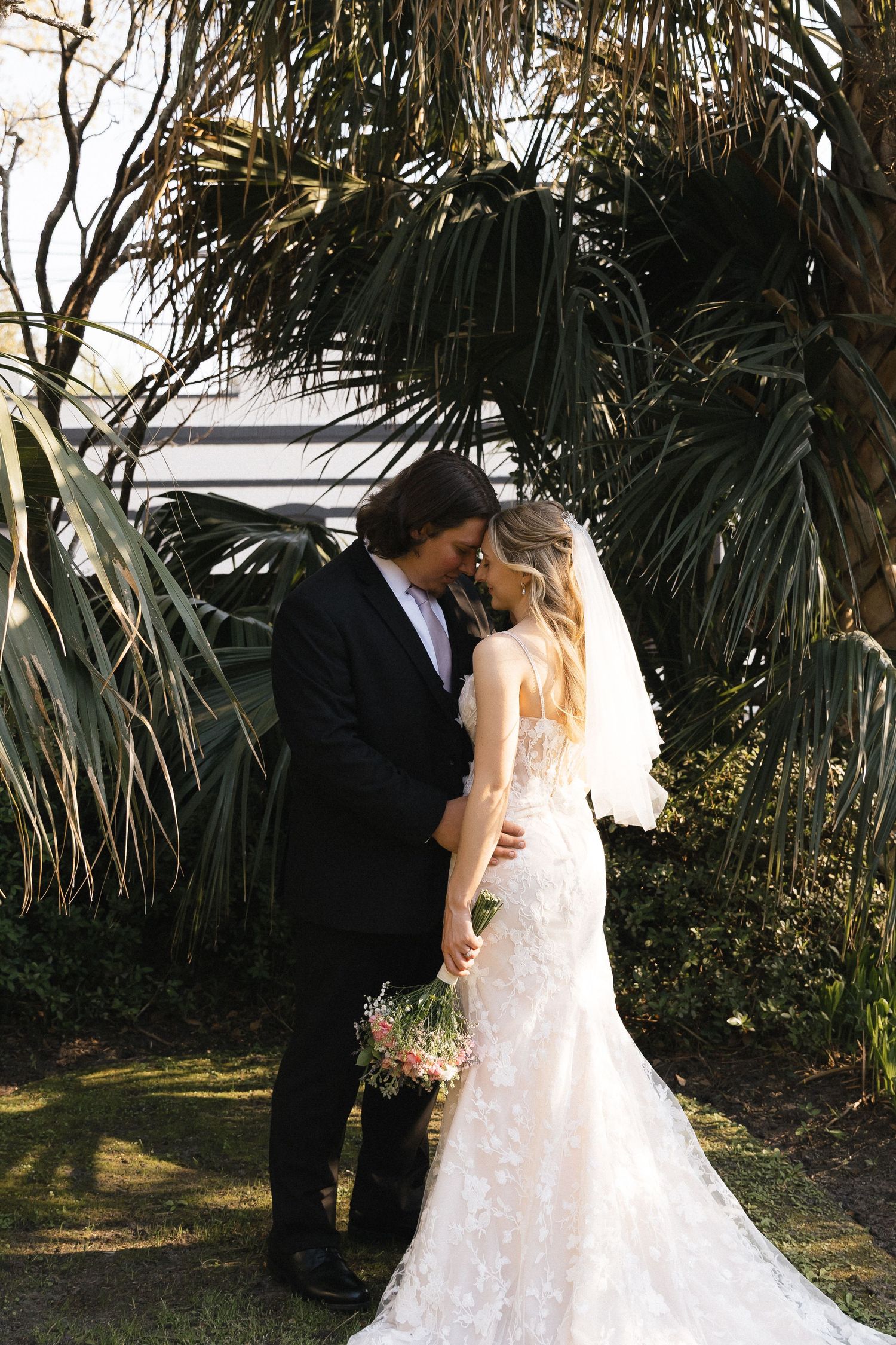 Bride and groom share a romantic forehead touch in a lush garden, bride wearing lace gown holding floral bouquet.