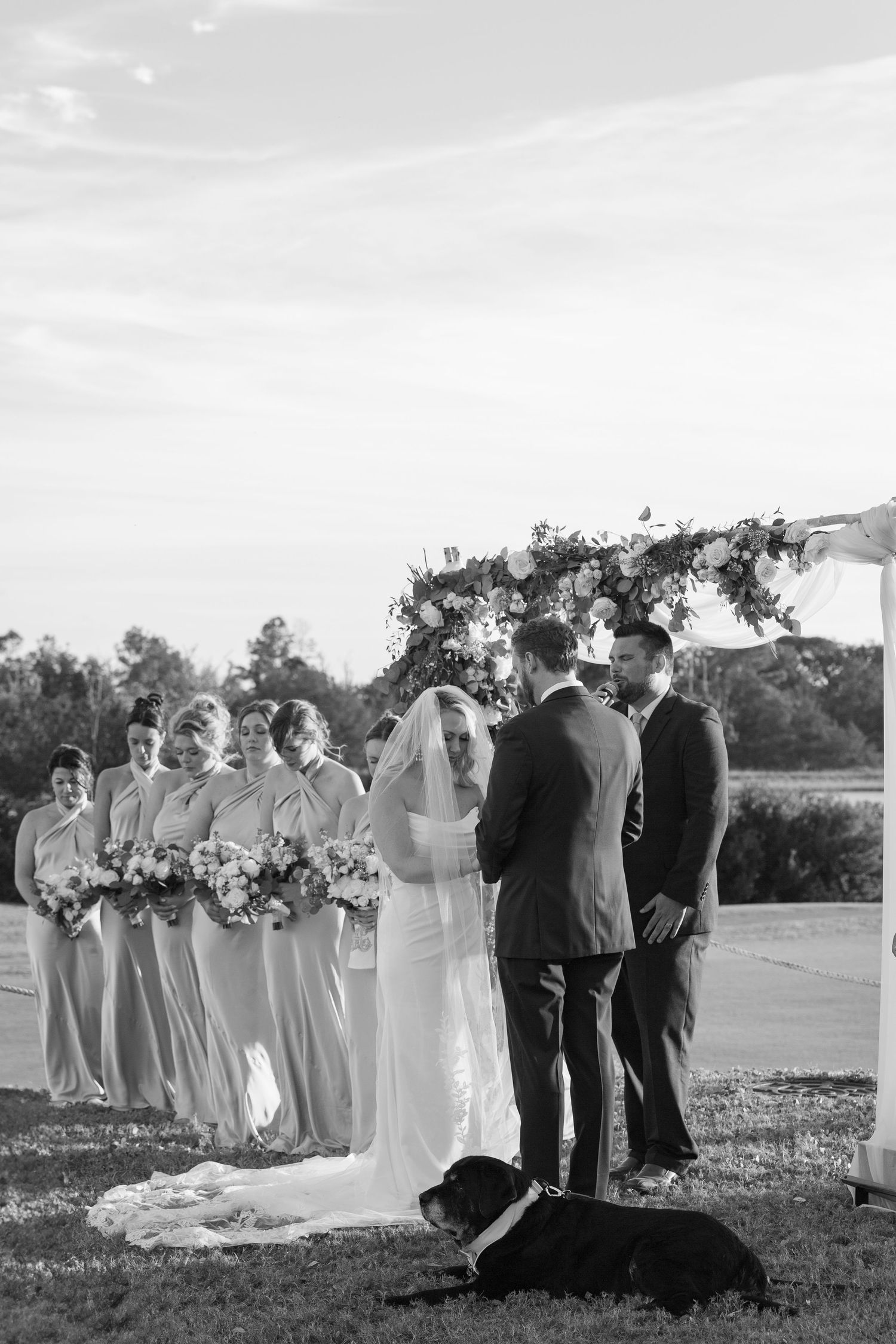 Black and white outdoor wedding ceremony with bride, groom, bridesmaids, and a dog lying at the altar.
