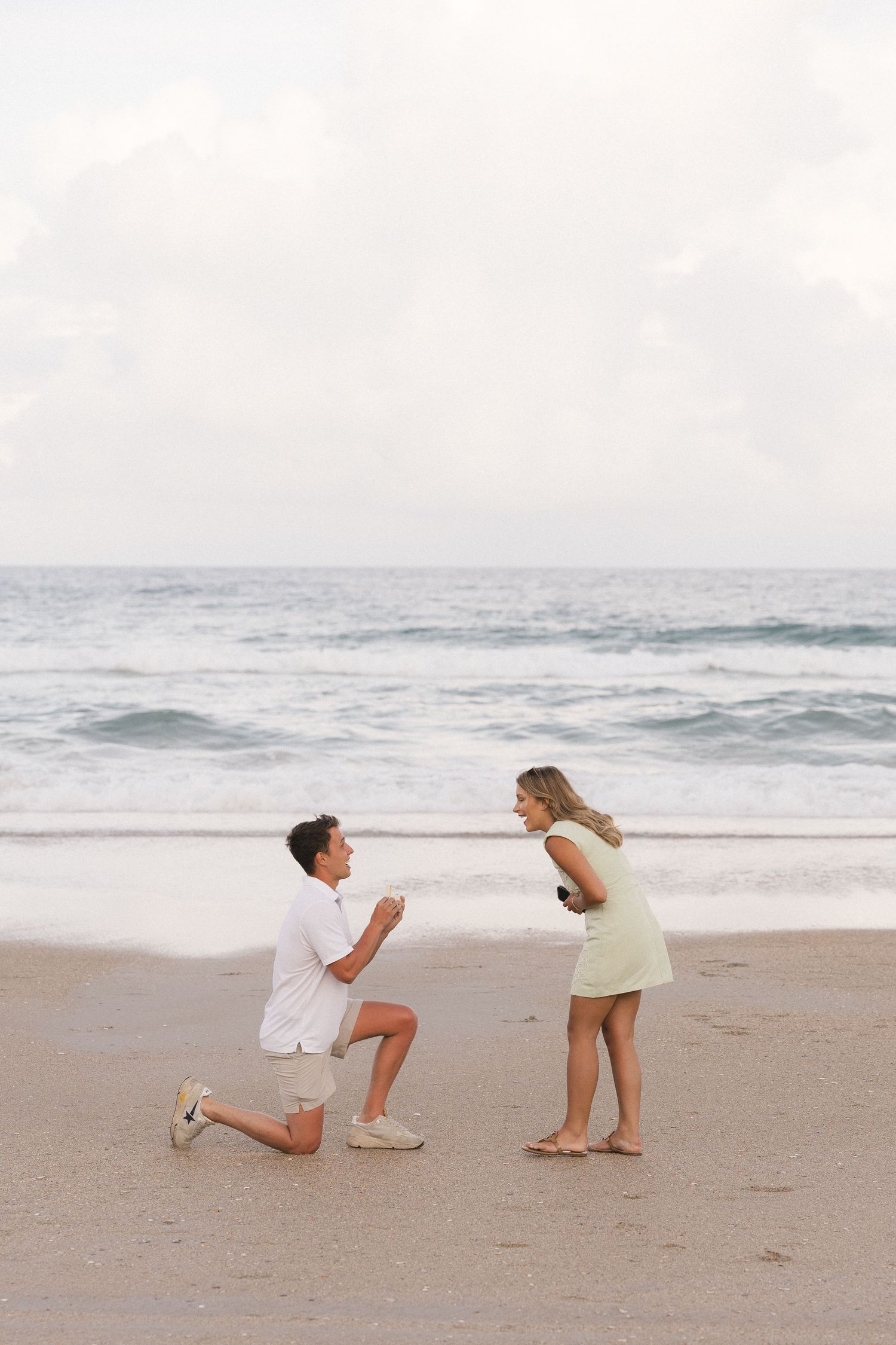 Man proposes to woman on one knee at a sandy beach with ocean waves in the background.