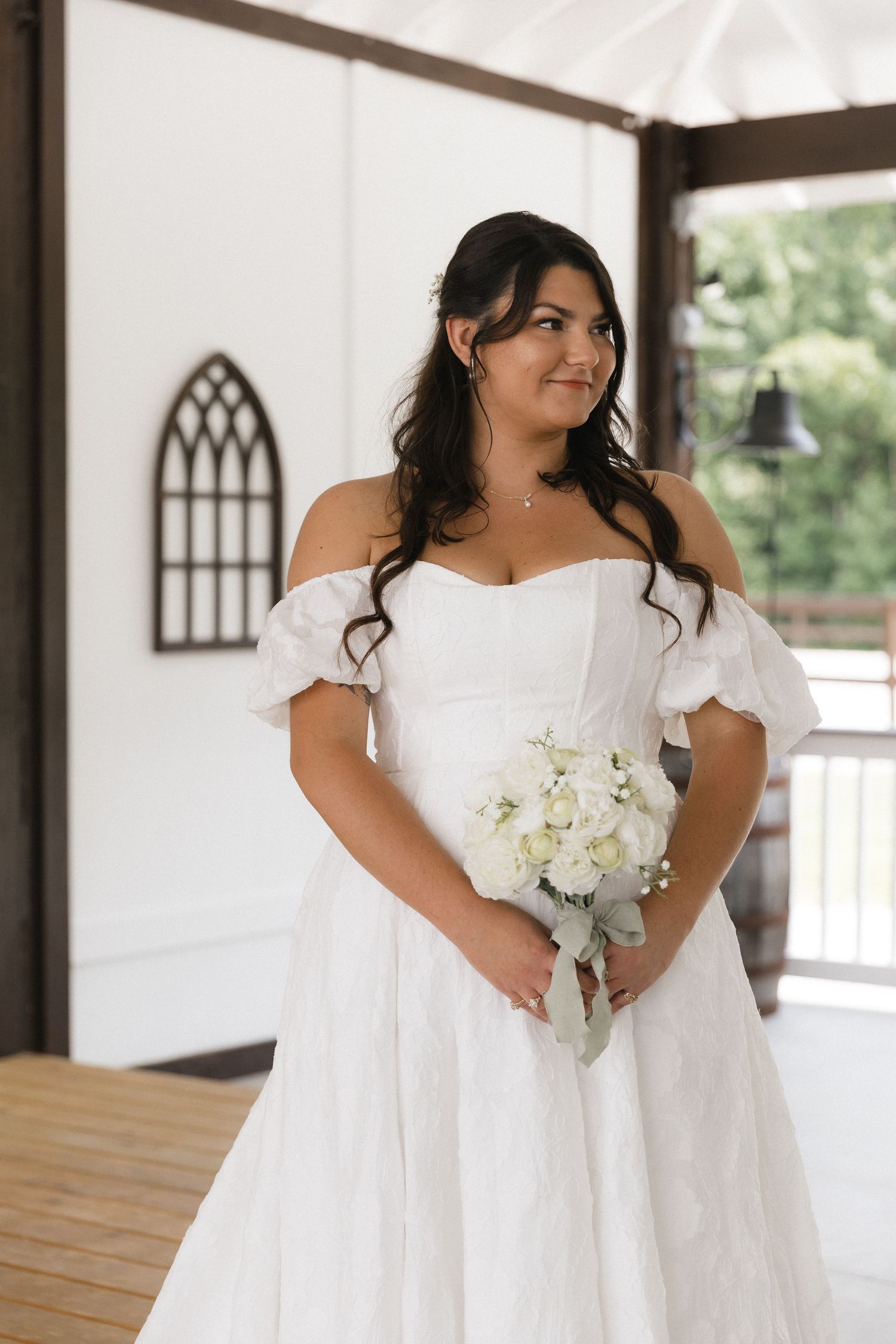 A bride in an off-shoulder white gown holds a bouquet of white roses, smiling softly inside a bright chapel.
