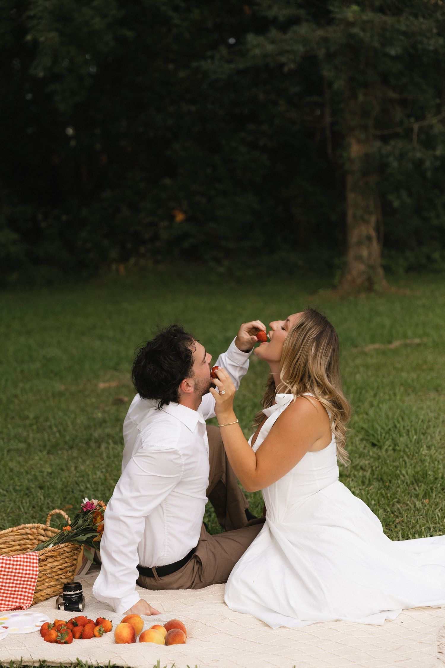 Couple in white attire sharing fruit during a romantic outdoor picnic on a blanket surrounded by peaches and a wicker basket.