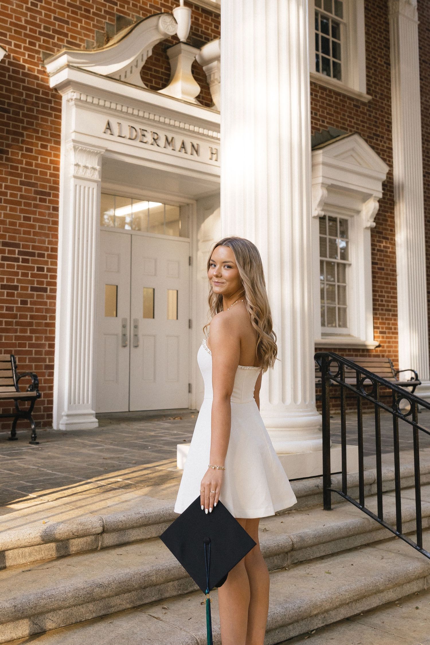 A young woman in a white dress holds a graduation cap on the steps of Alderman Hall at a university.