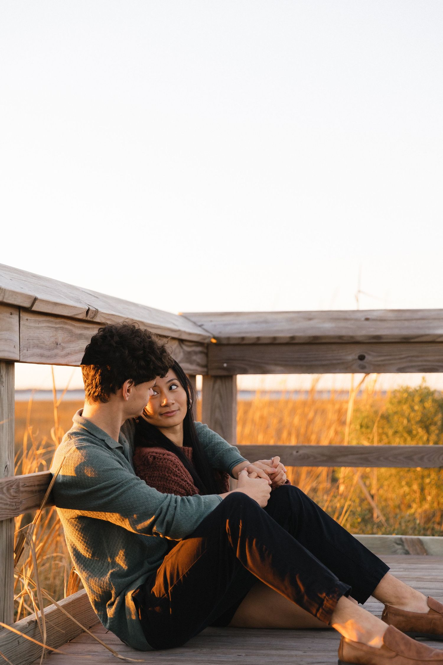 Couple sitting together on a wooden deck at golden hour, holding hands and enjoying a scenic marshland sunset.