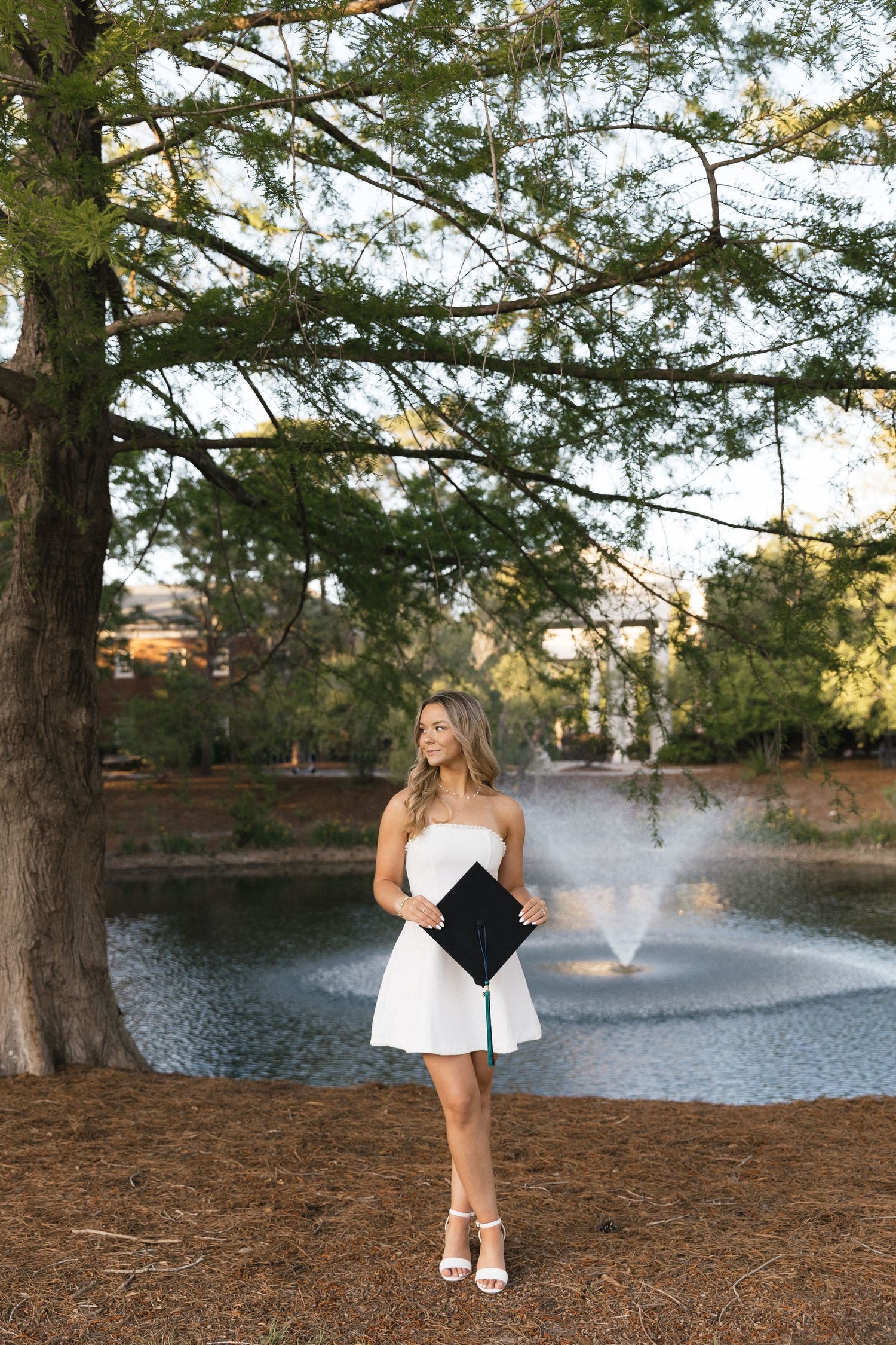 Young woman in white dress holding graduation cap by a pond under large trees.