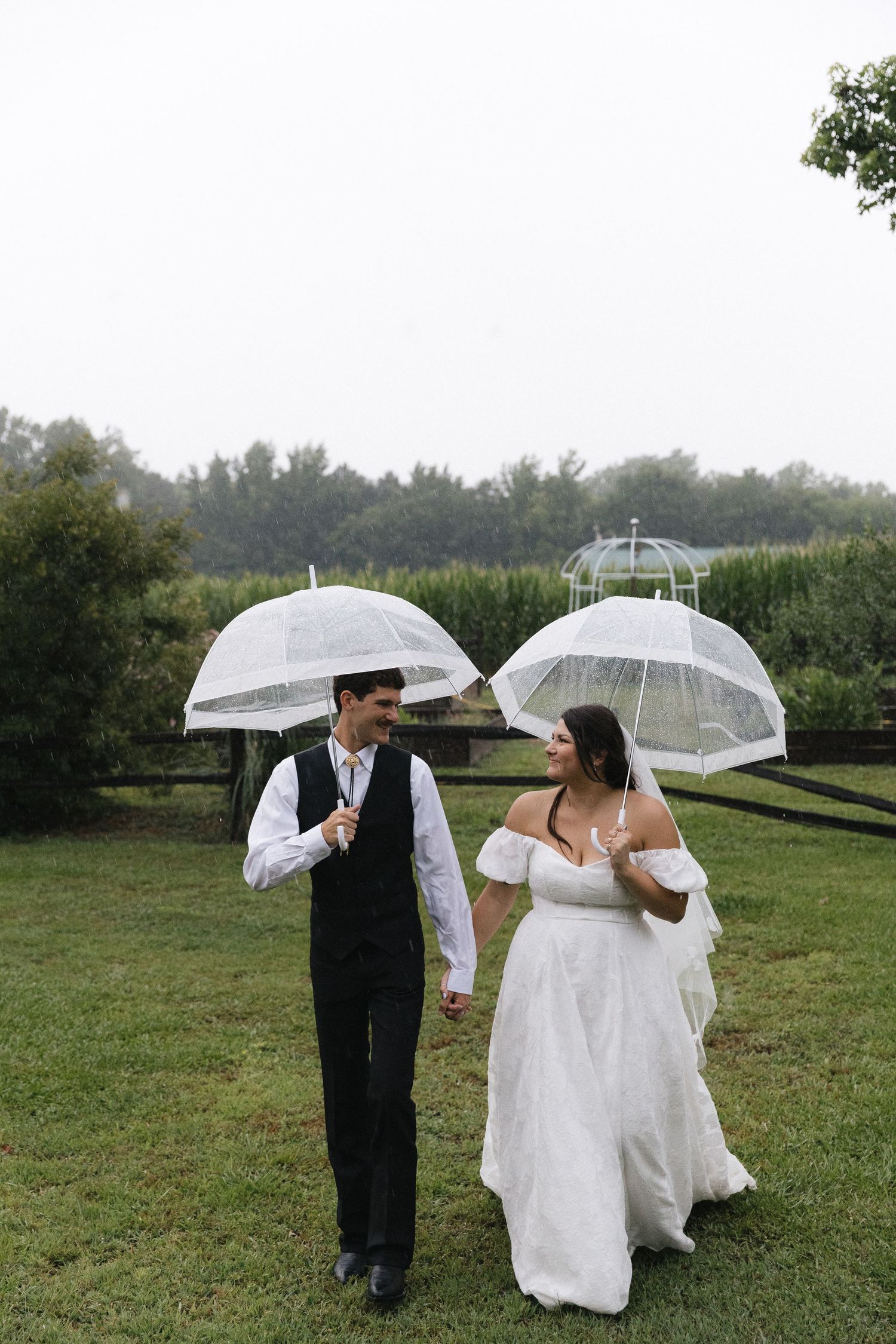 A bride and groom walk hand in hand on a rainy wedding day, each holding clear dome umbrellas outdoors.