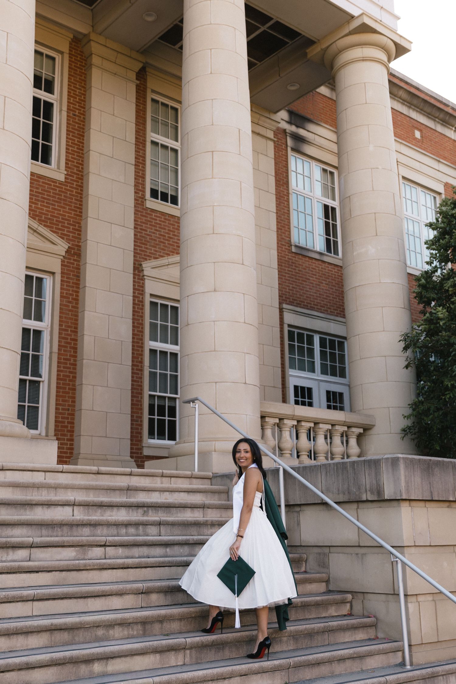 Graduate in white dress holding green cap on university building steps with large columns.