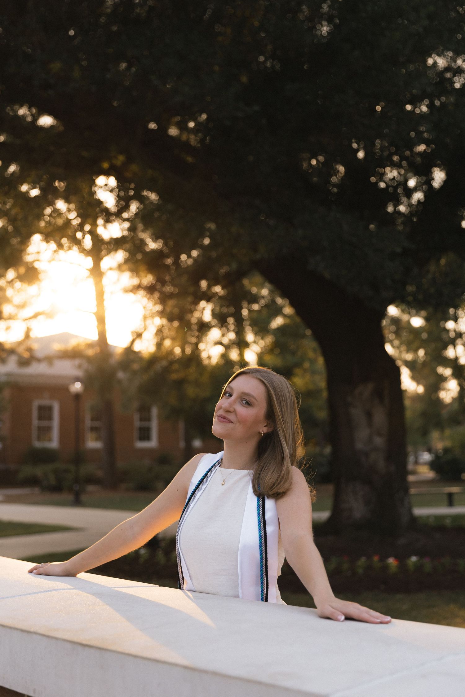 Young woman in white dress leans on stone wall at golden hour, with trees and brick building in background.