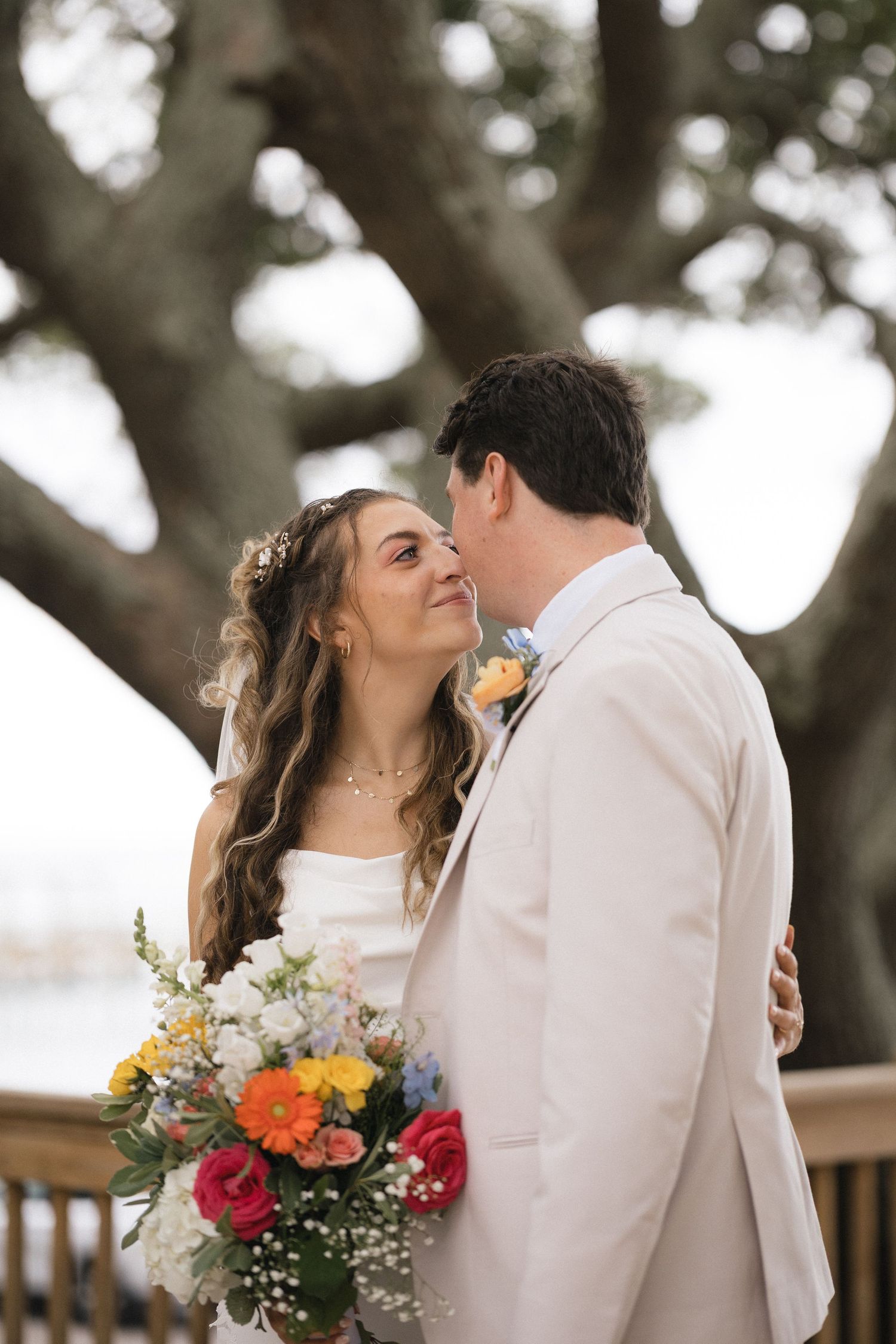A bride holding a colorful bouquet shares a tender moment with her groom in a white suit beneath a large oak tree.