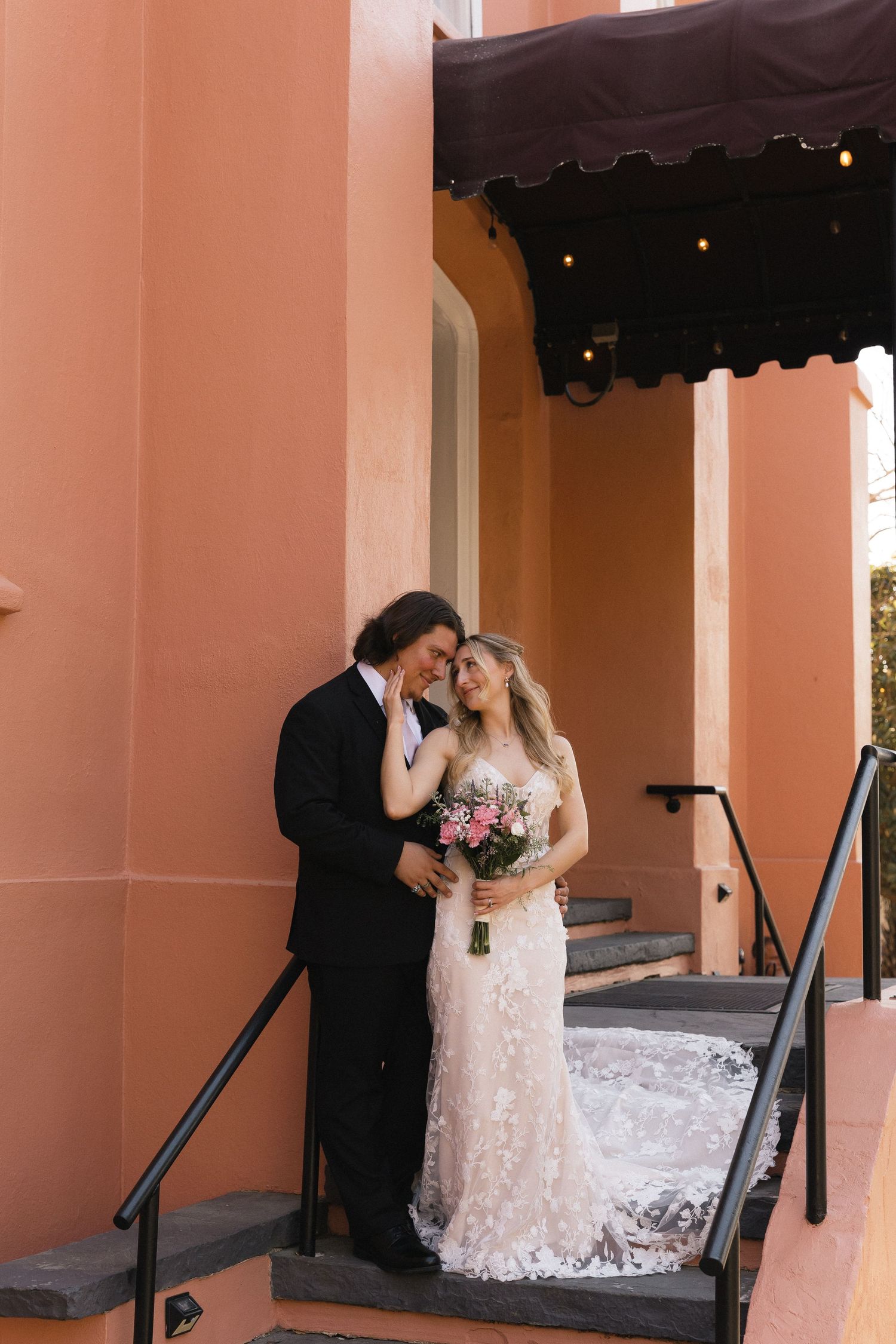 Bride and groom share a romantic moment on steps outside a coral building, bride holding a pink floral bouquet.
