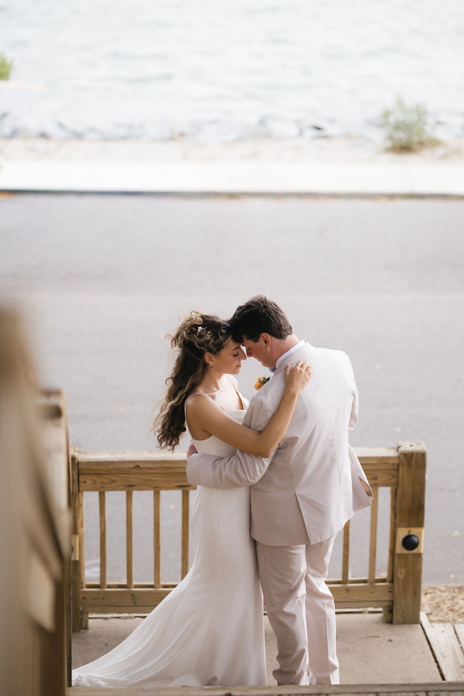 Bride and groom share a romantic moment on a waterfront deck, dressed in white wedding attire.