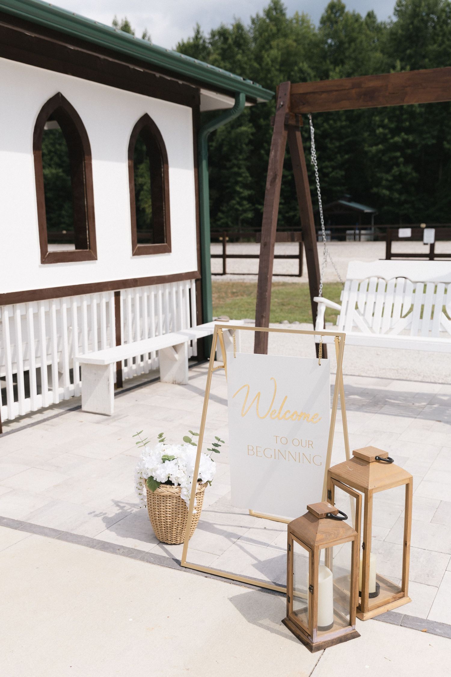 Elegant wedding welcome sign on gold easel with lanterns and flowers outside a white venue.
