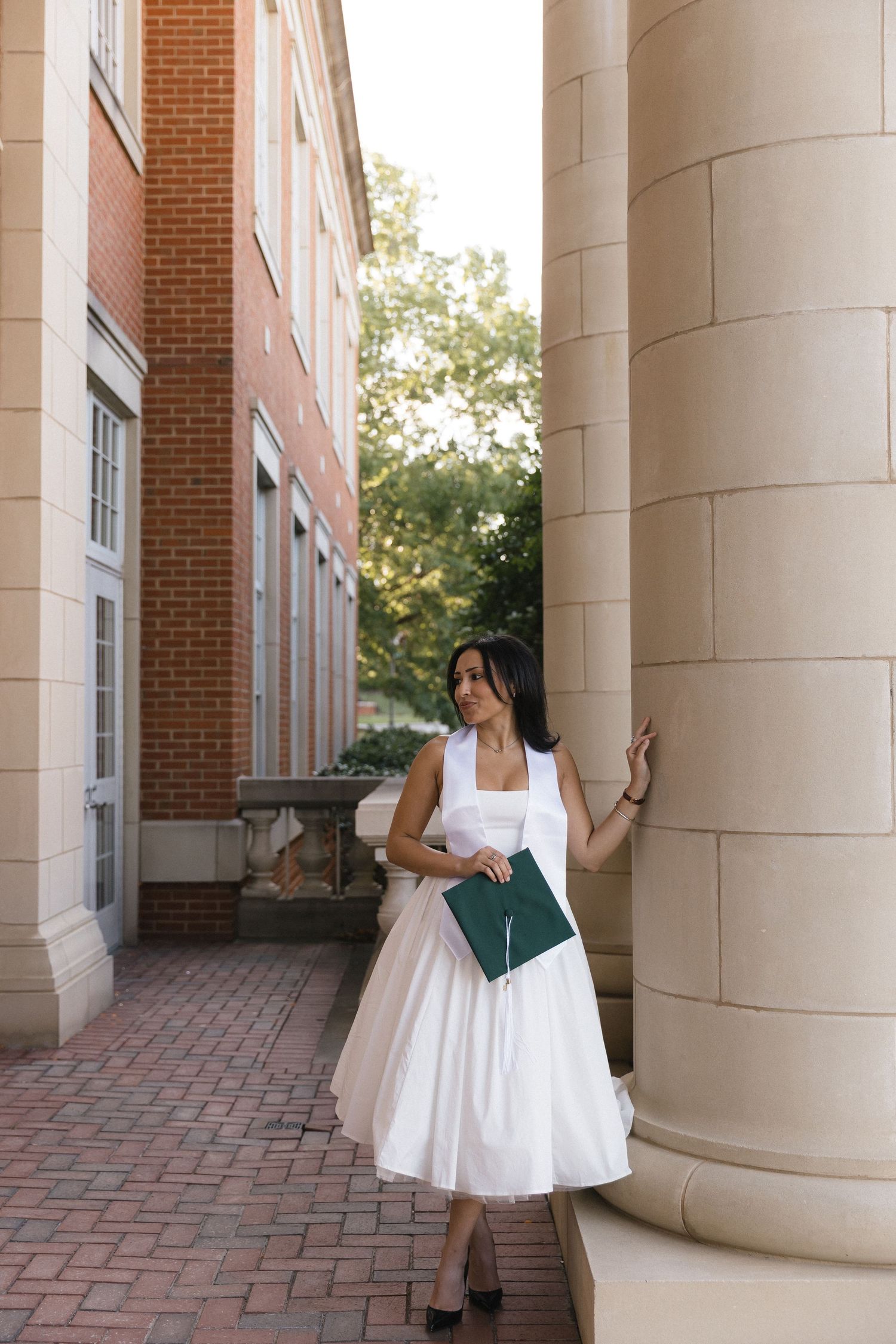 Woman in white dress holding green diploma folder, posing by stone columns of a brick university building.