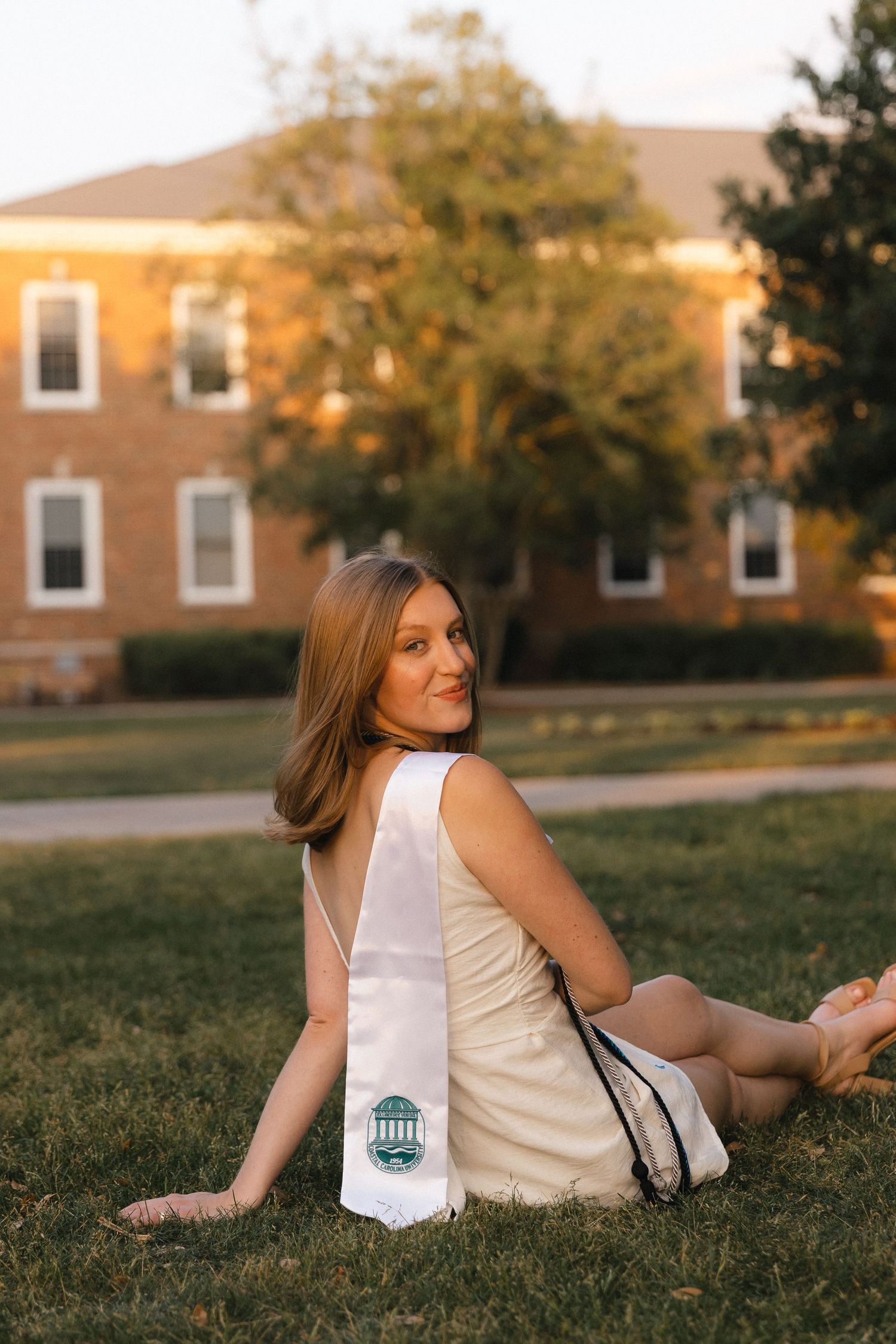 A young woman in a white dress with a graduation stole sits on grass, smiling over her shoulder near a brick building.