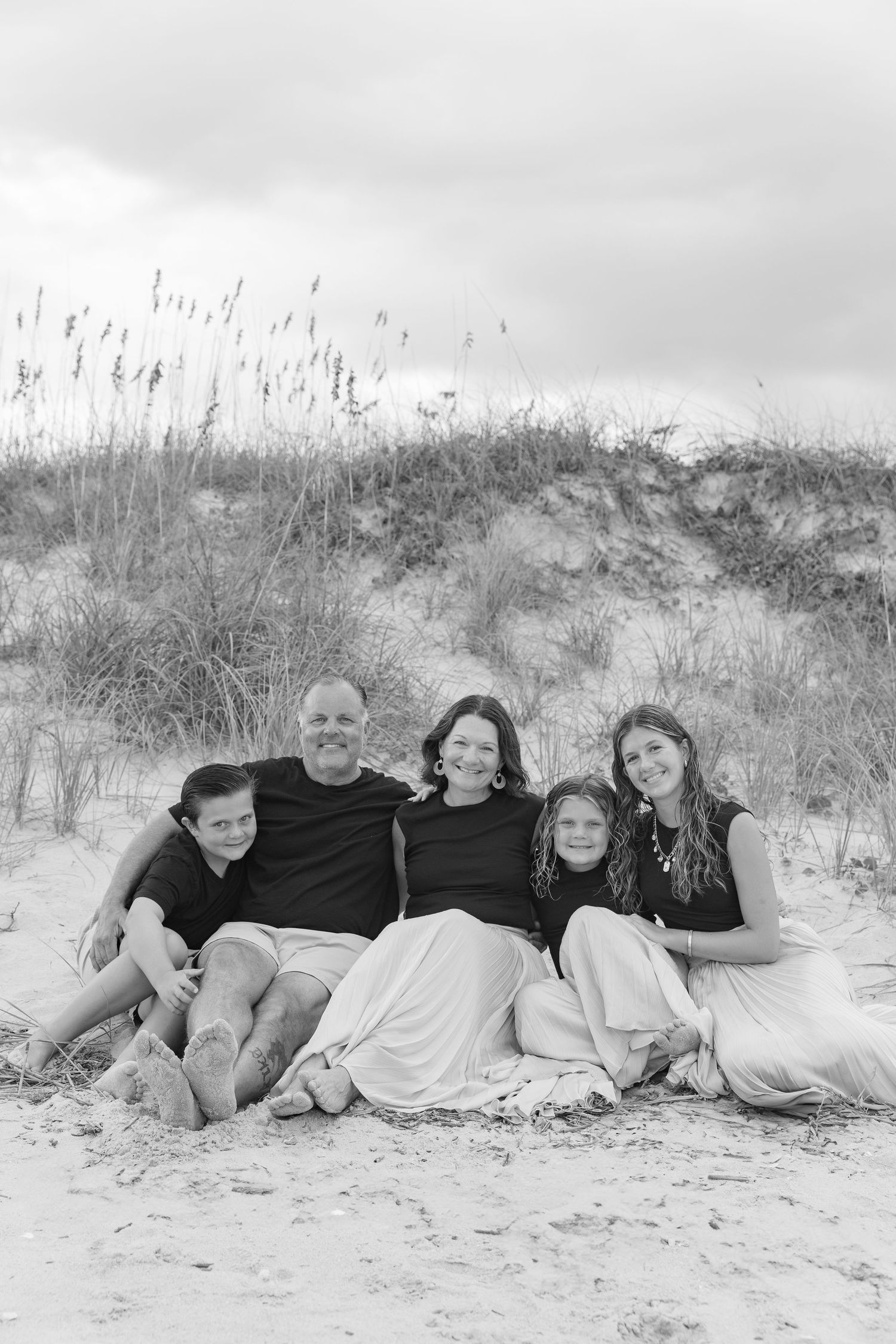Black and white family portrait of five people sitting together on a sandy beach with sea oats and dunes in background.