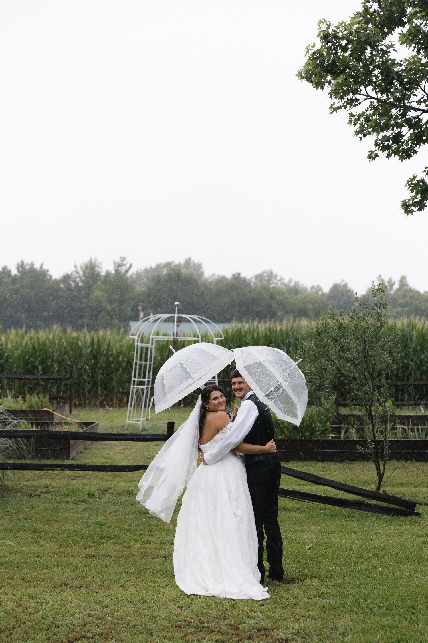 Bride and groom sharing a romantic moment under clear umbrellas in the rain on their wedding day outdoors.