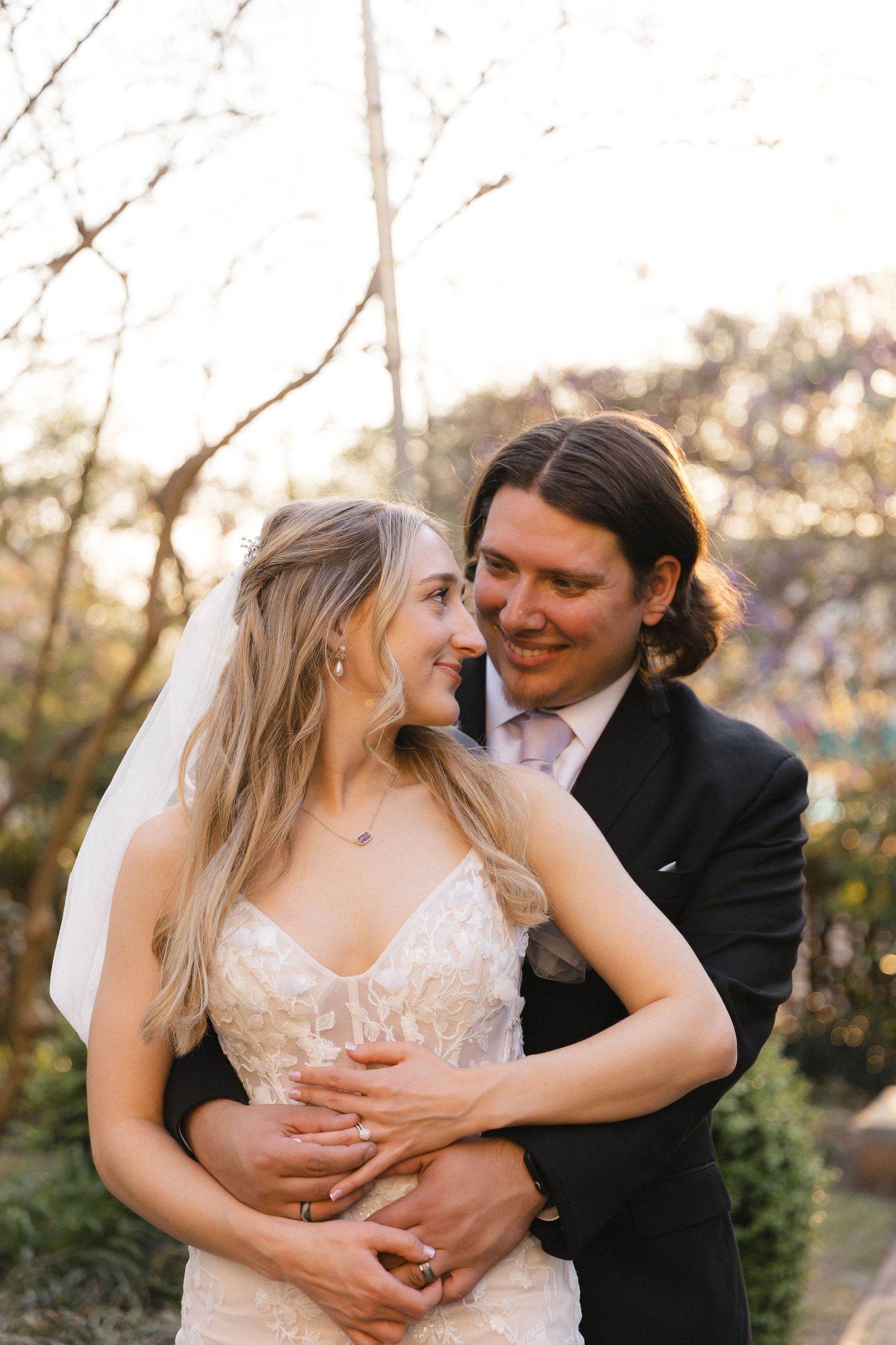A groom embraces his bride from behind in a sunlit garden, both smiling warmly in wedding attire.