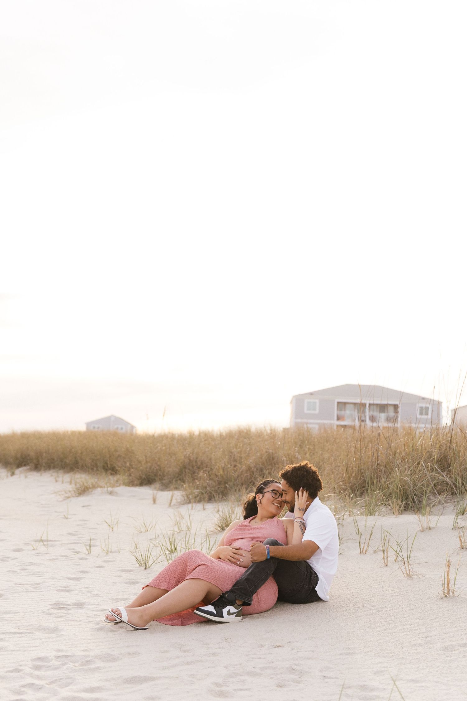 Couple sitting on sandy beach dunes at sunset, woman in pink dress with baby bump, man in white shirt embracing her.