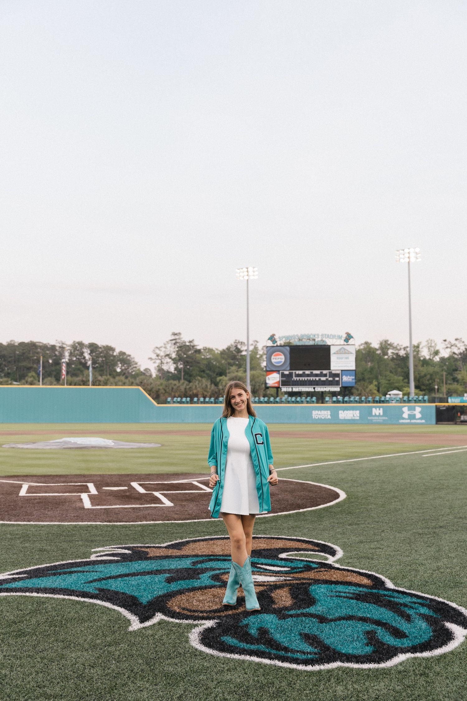 Graduate in teal cap and gown stands on Coastal Carolina Chanticleers baseball field logo in white dress.