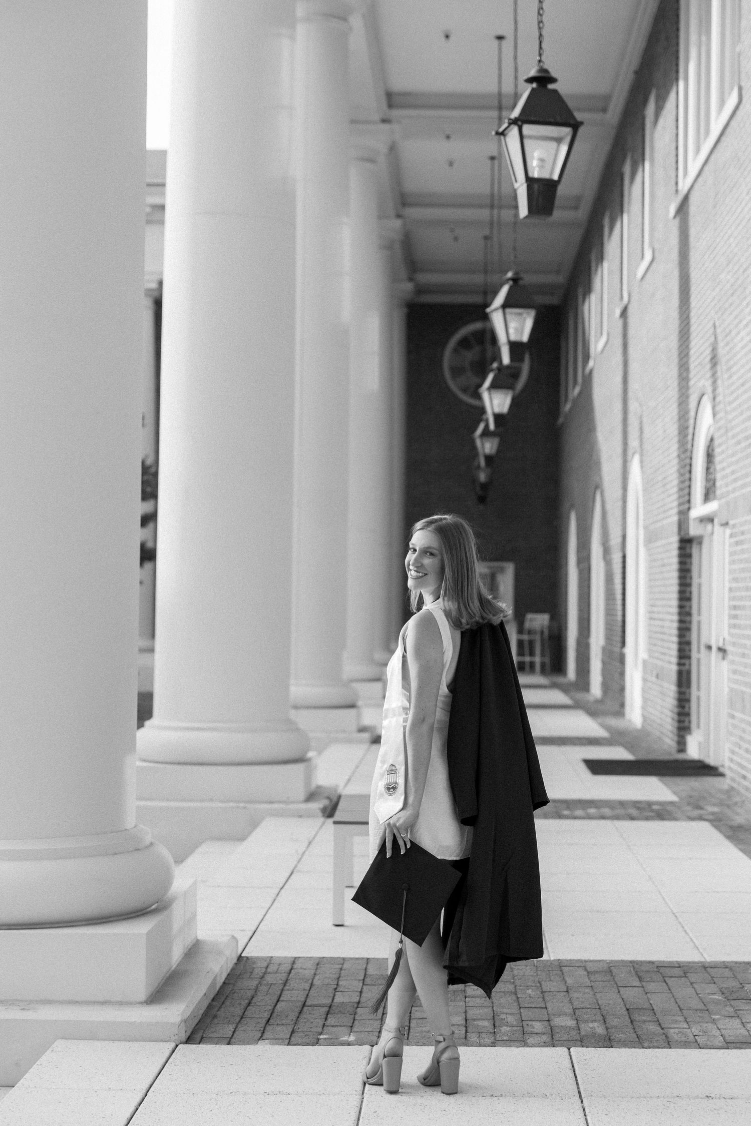 Black and white photo of a smiling woman in a graduation gown walking along a columned corridor.