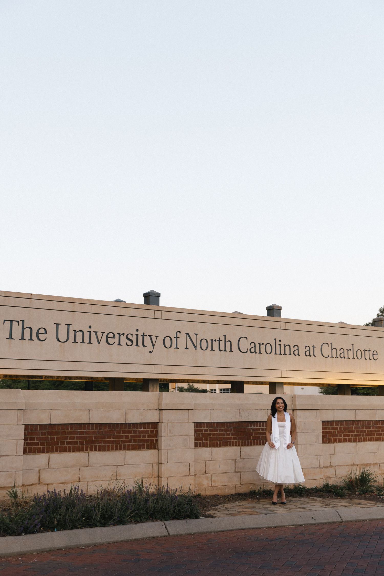 Graduate in white dress posing by University of North Carolina at Charlotte campus sign.