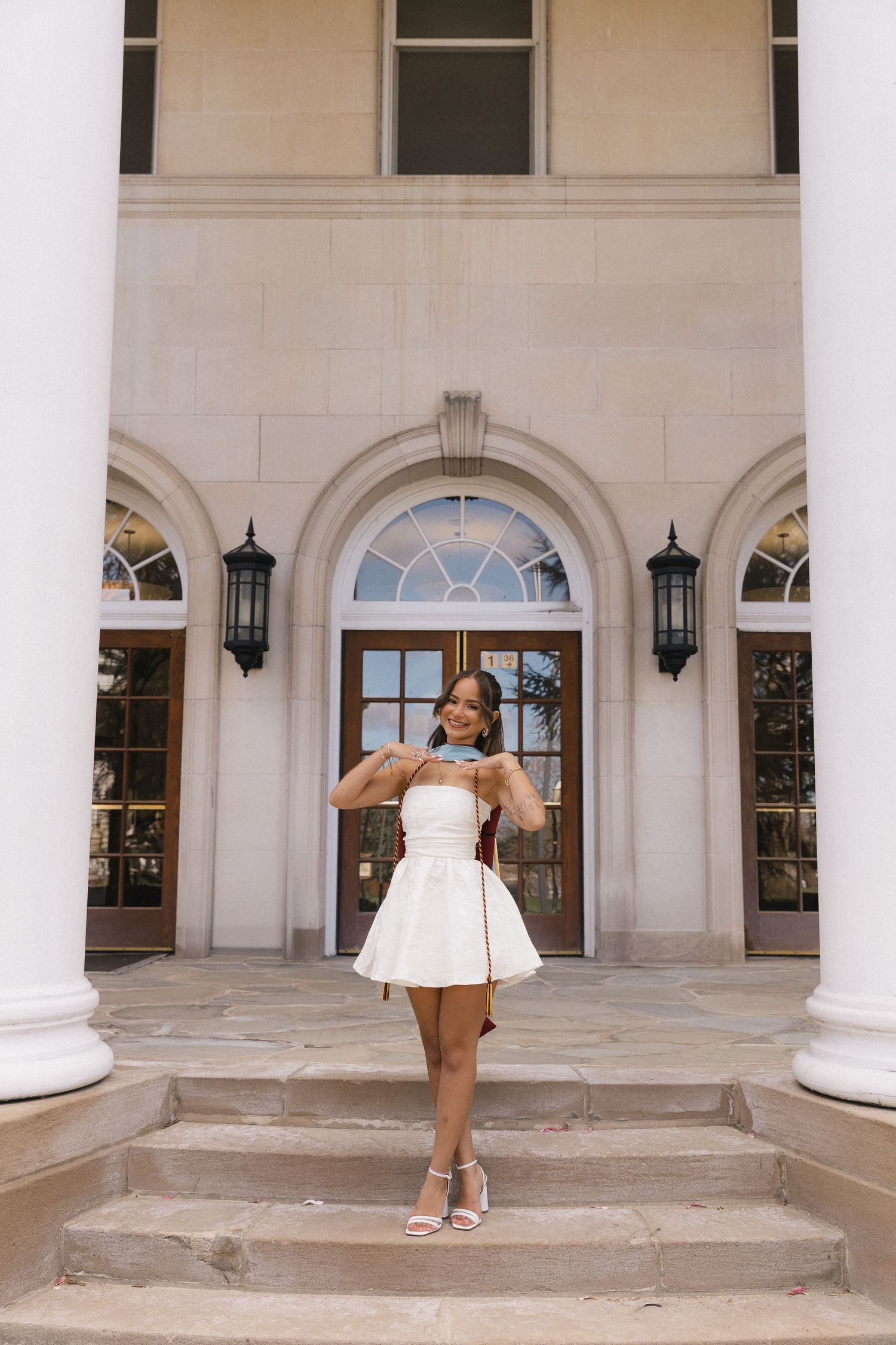 Young woman in white mini dress posing on steps of elegant building with arched doorways.