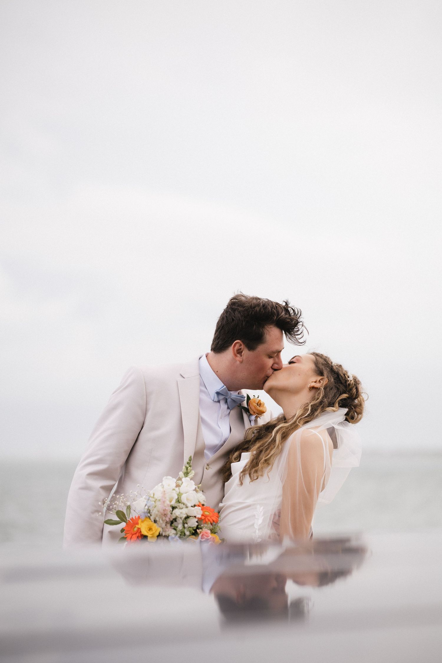Bride and groom share a romantic kiss on a beach, she holds colorful bouquet, he wears tan suit with blue bow tie.