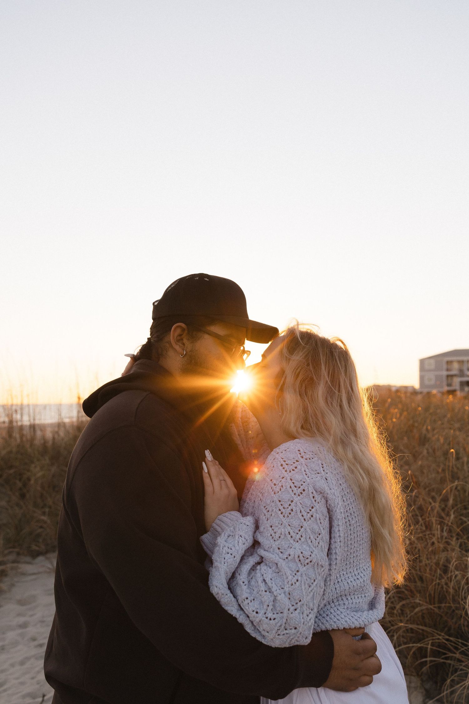 Couple sharing a romantic sunset kiss on a beach, surrounded by golden sea grass and warm sunlight.