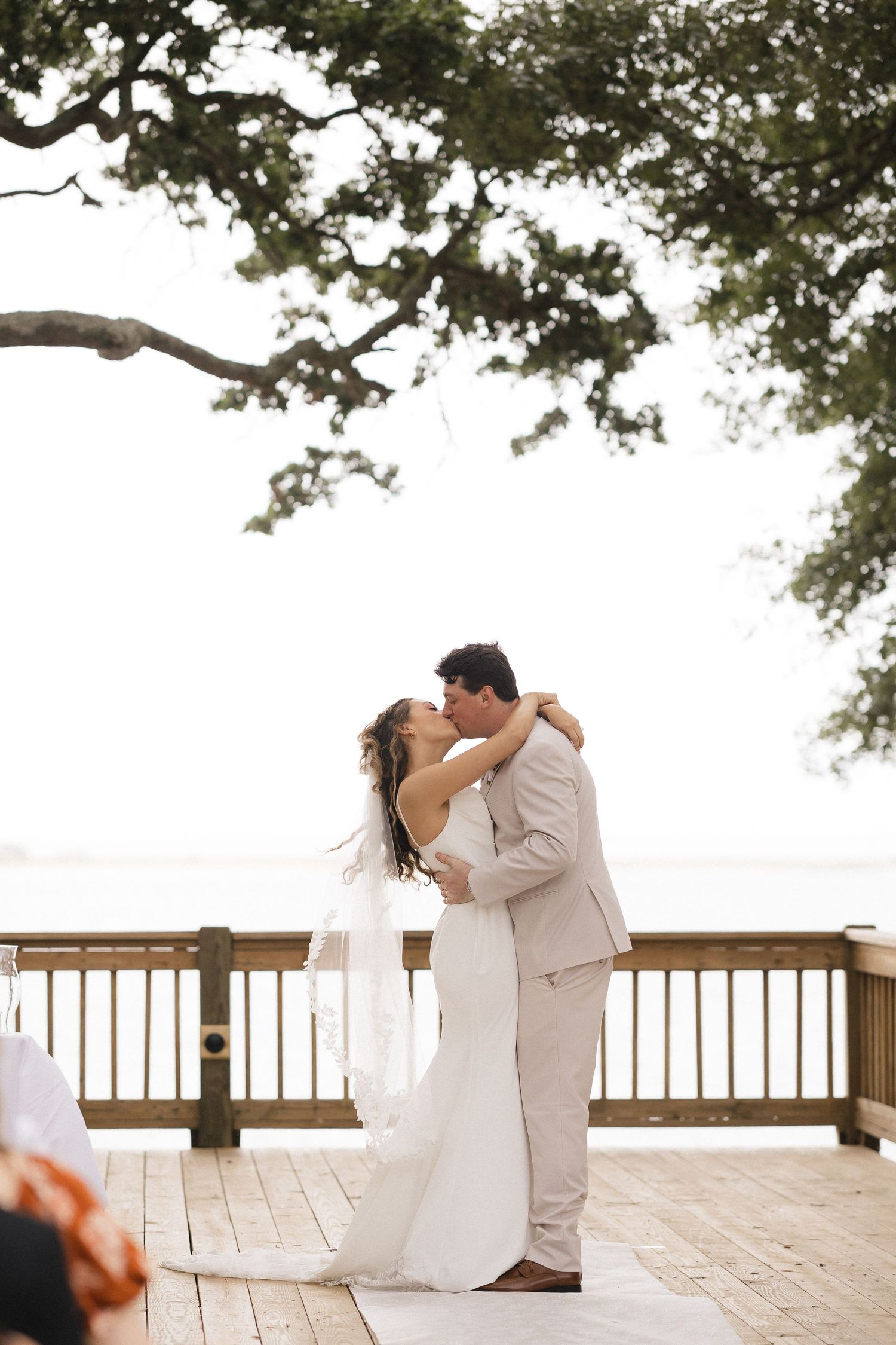 Bride and groom share first kiss on wooden deck overlooking water, surrounded by oak trees at outdoor wedding ceremony.