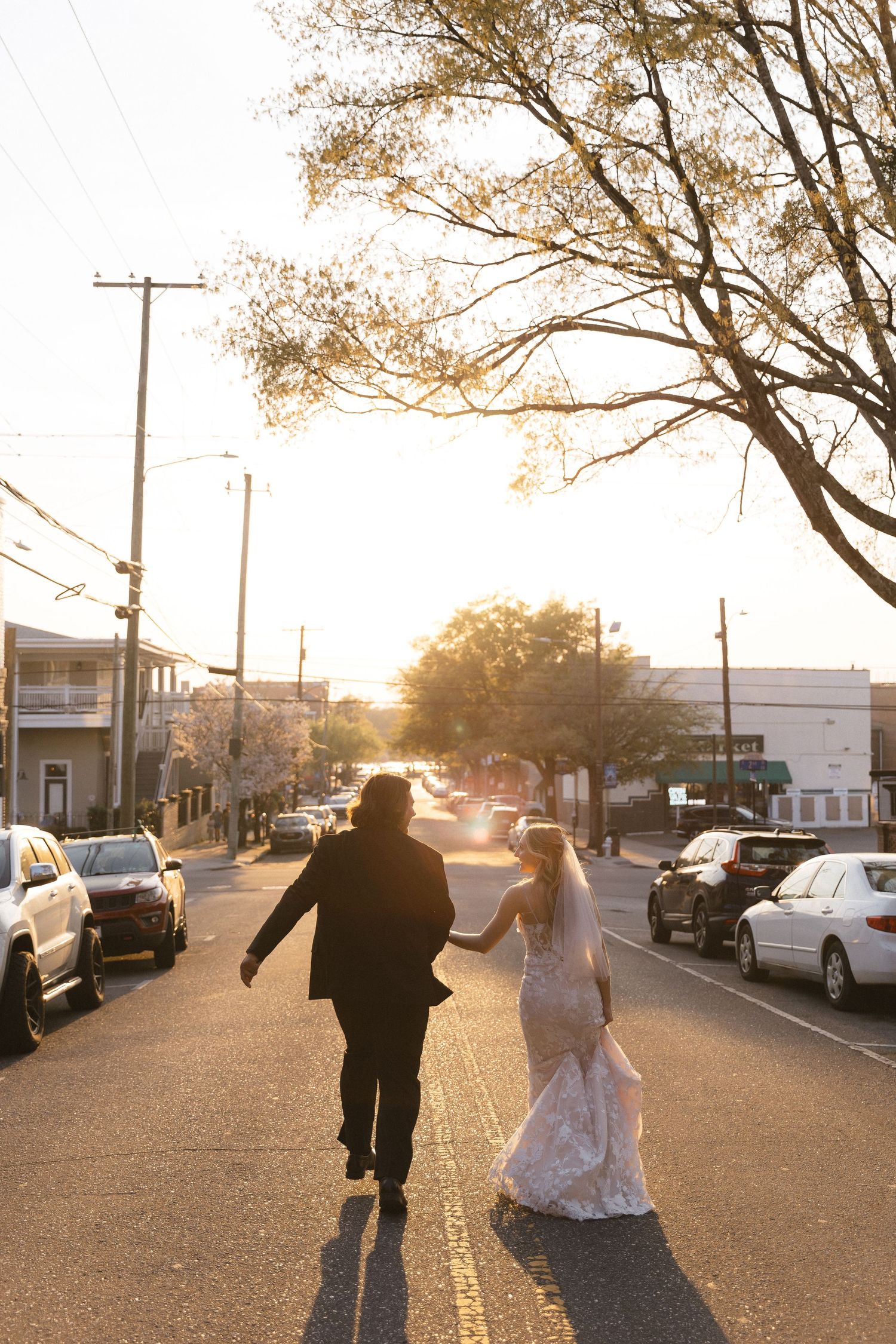 Bride and groom walk hand-in-hand down a sunlit street at golden hour, cars and trees lining the road.