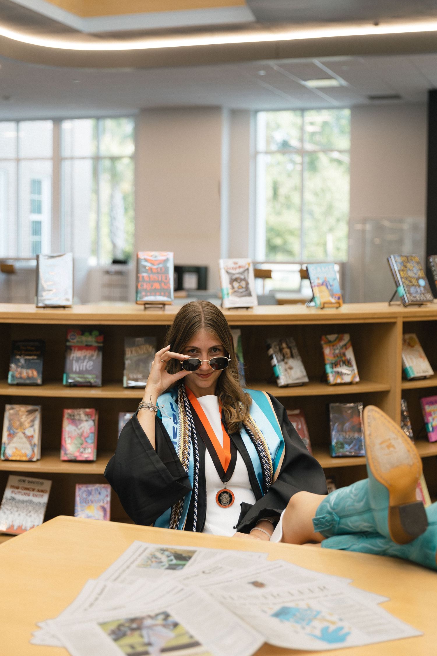 Graduate in black gown and teal sashes poses with sunglasses in a school library setting.