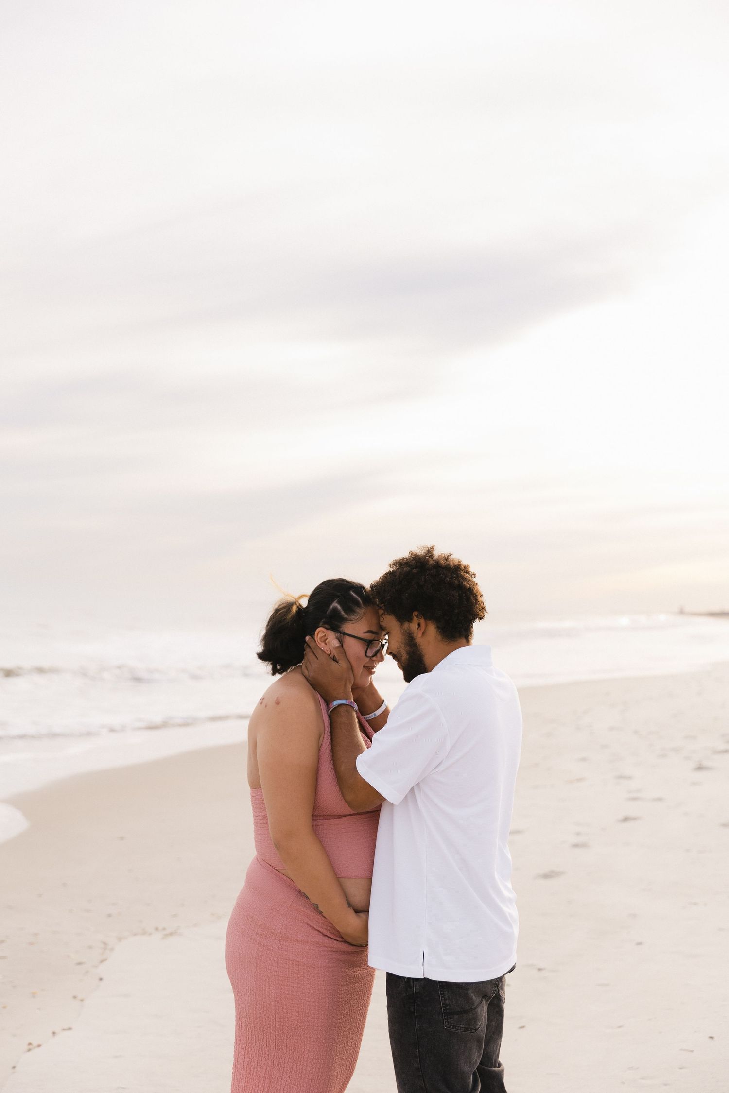 Couple sharing an intimate moment on a sandy beach at sunset, woman in pink dress with visible baby bump.