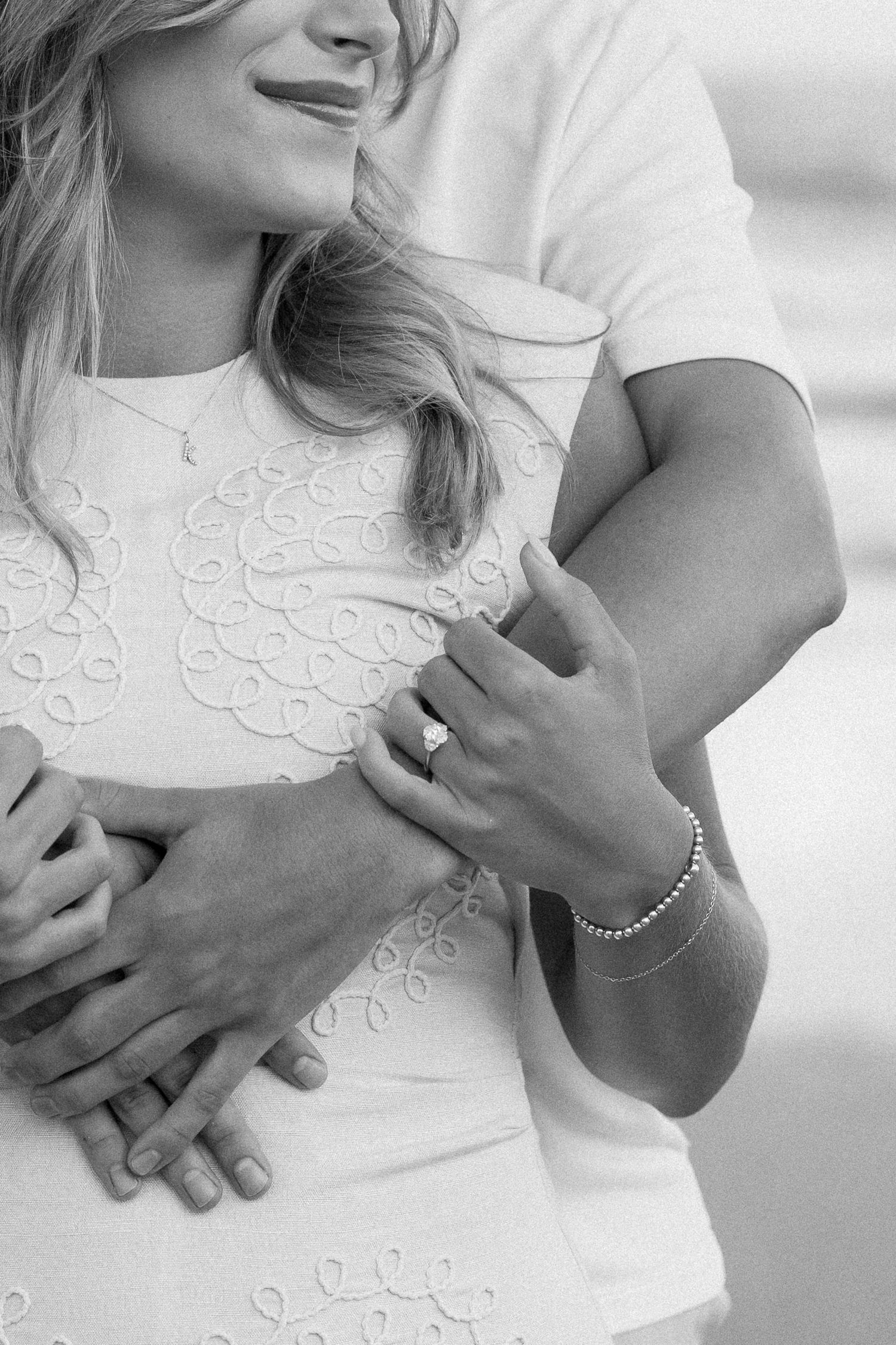 Black and white photo of a couple embracing, woman wearing an engagement ring and pearl bracelet.