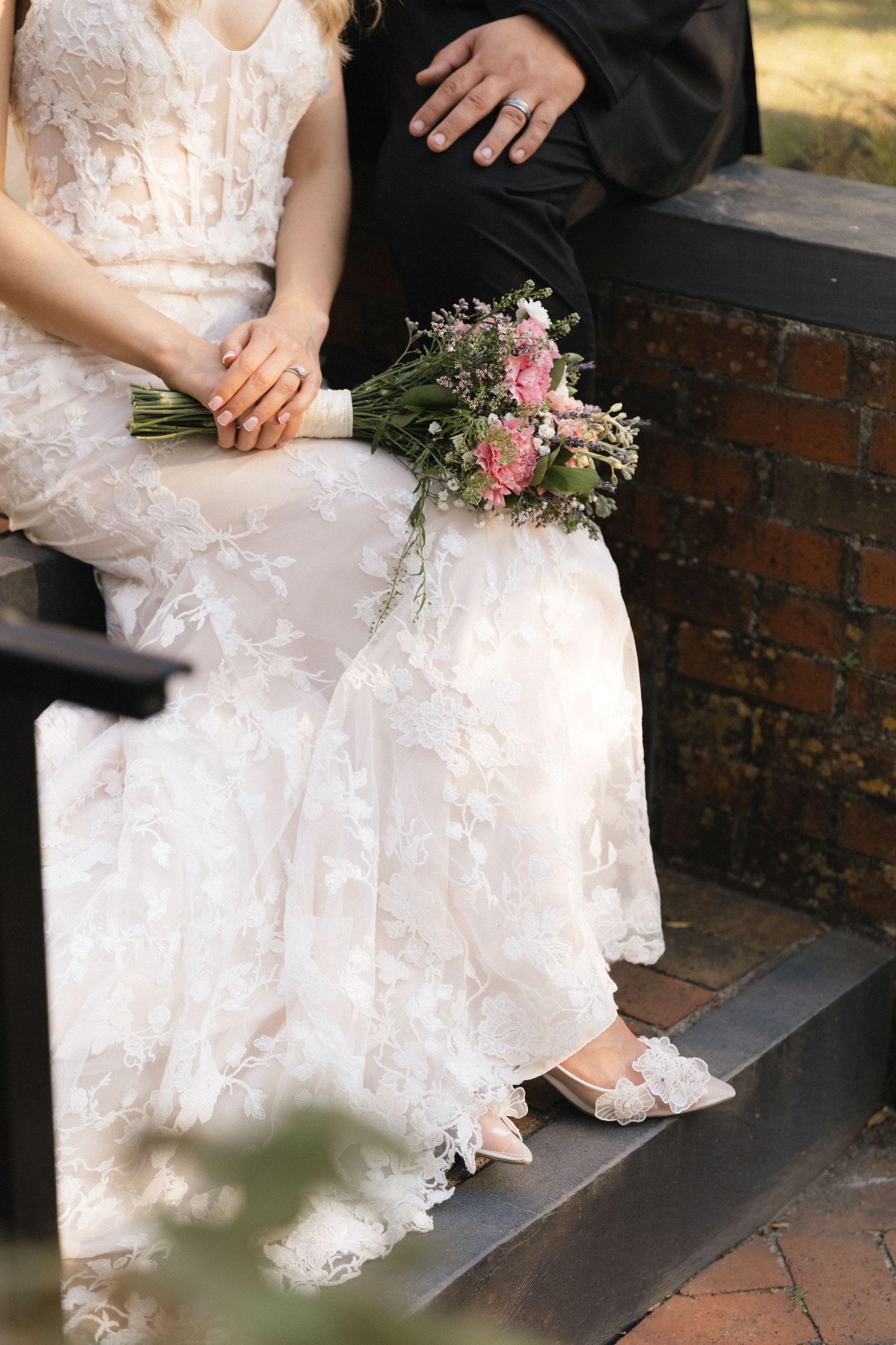 Bride in lace gown holding pink floral bouquet seated beside groom in black suit on brick steps.